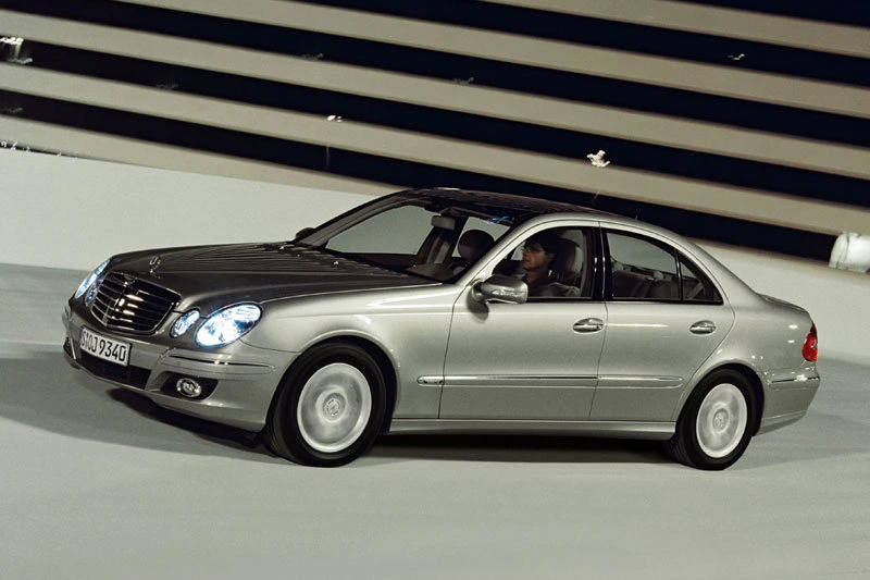 Silberner Mercedes-Benz Limousine fährt auf einer modernen Straße, Seitenansicht, mit eingeschaltetem Licht und Fahrer am Steuer.