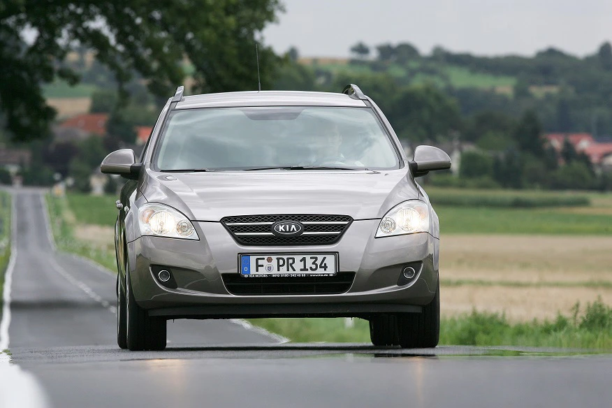 Silberner Kia Ceed fährt auf einer Landstraße durch eine grüne Landschaft, Bäume und Felder im Hintergrund.