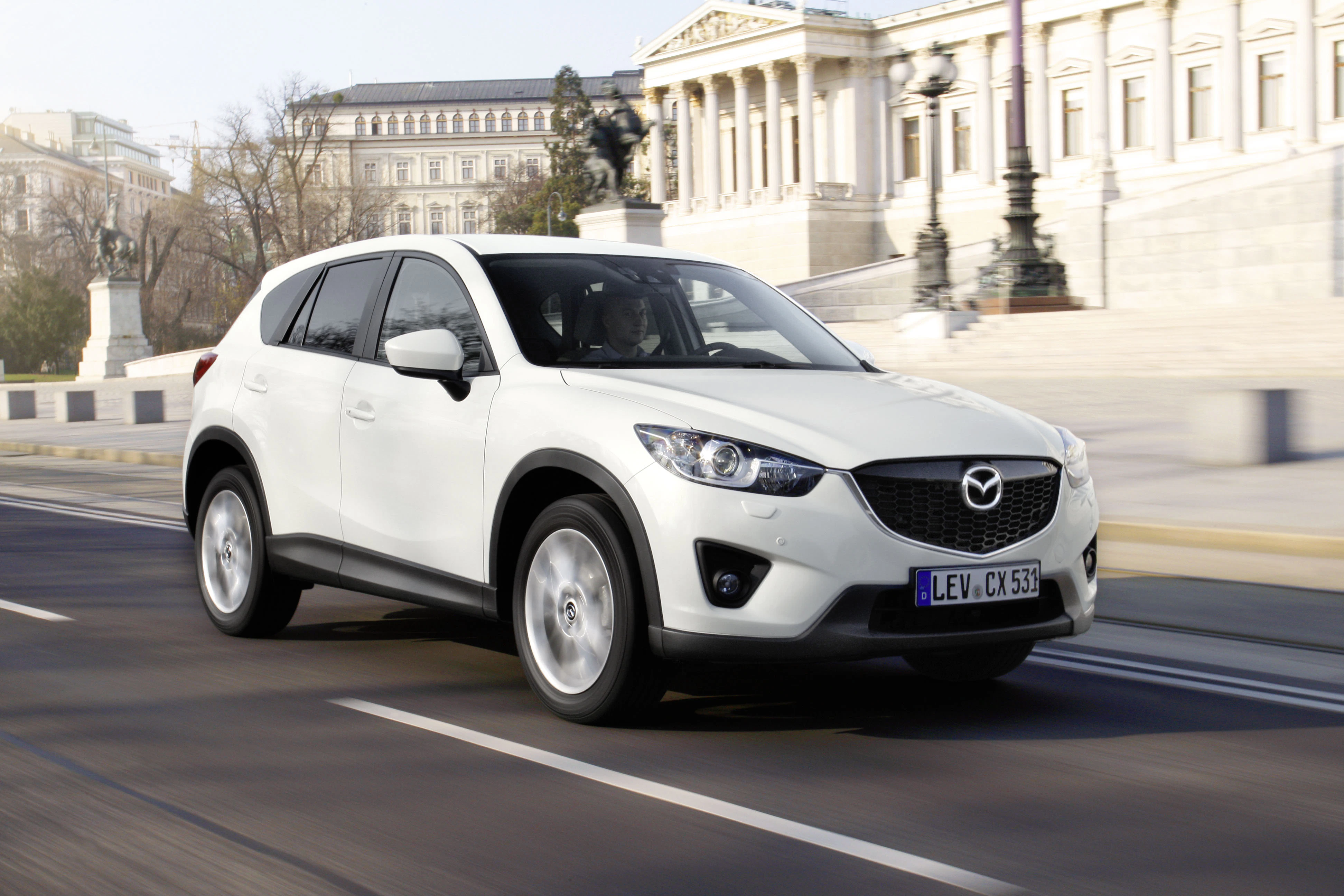 Weißer SUV, Mazda CX-5, fährt auf einer städtischen Straße mit historischen Gebäuden im Hintergrund.
