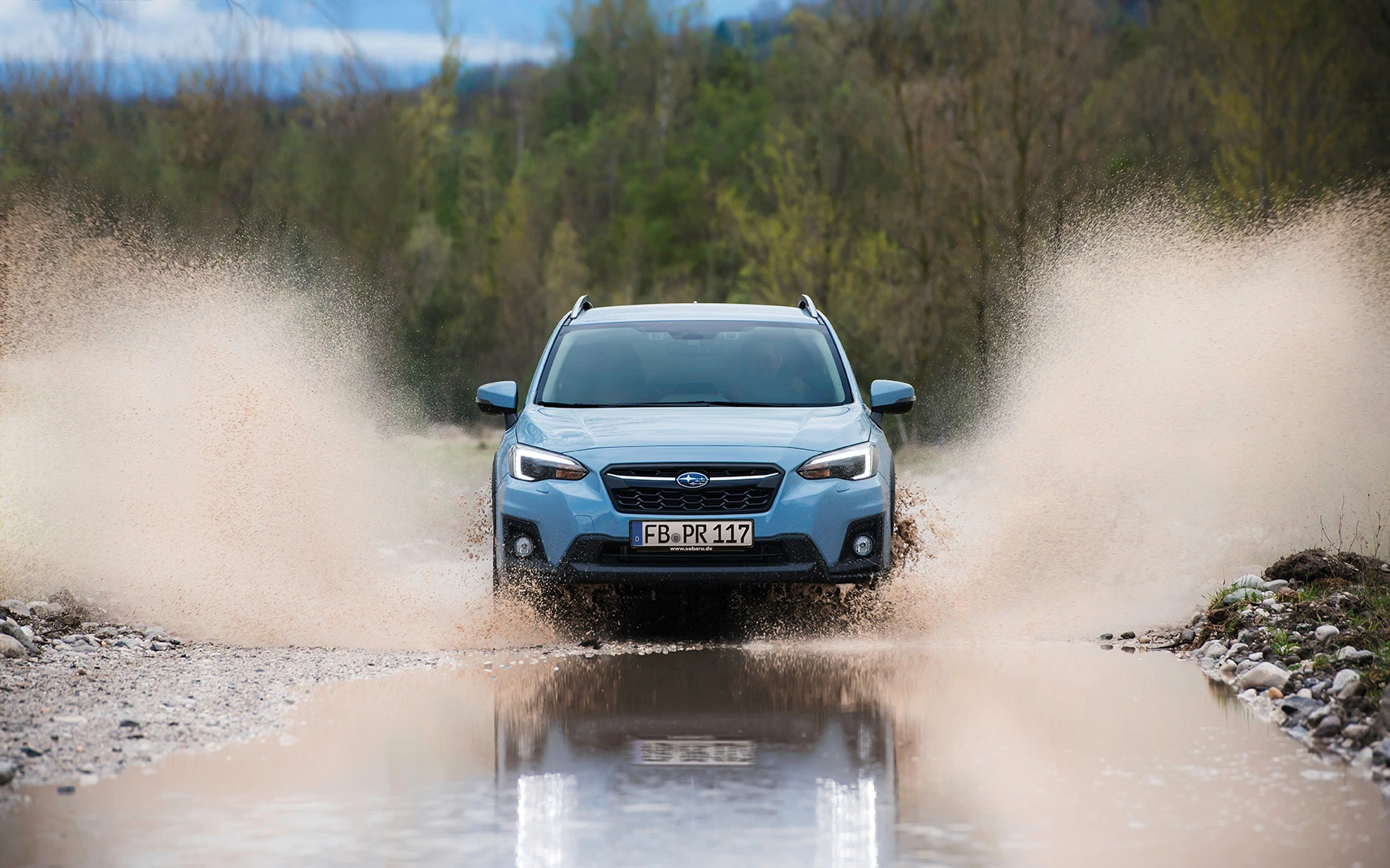 Ein blauer SUV fährt durch eine Pfütze auf einer Schotterstraße, spritzt Wasser seitlich auf, umgeben von Bäumen im Hintergrund.
