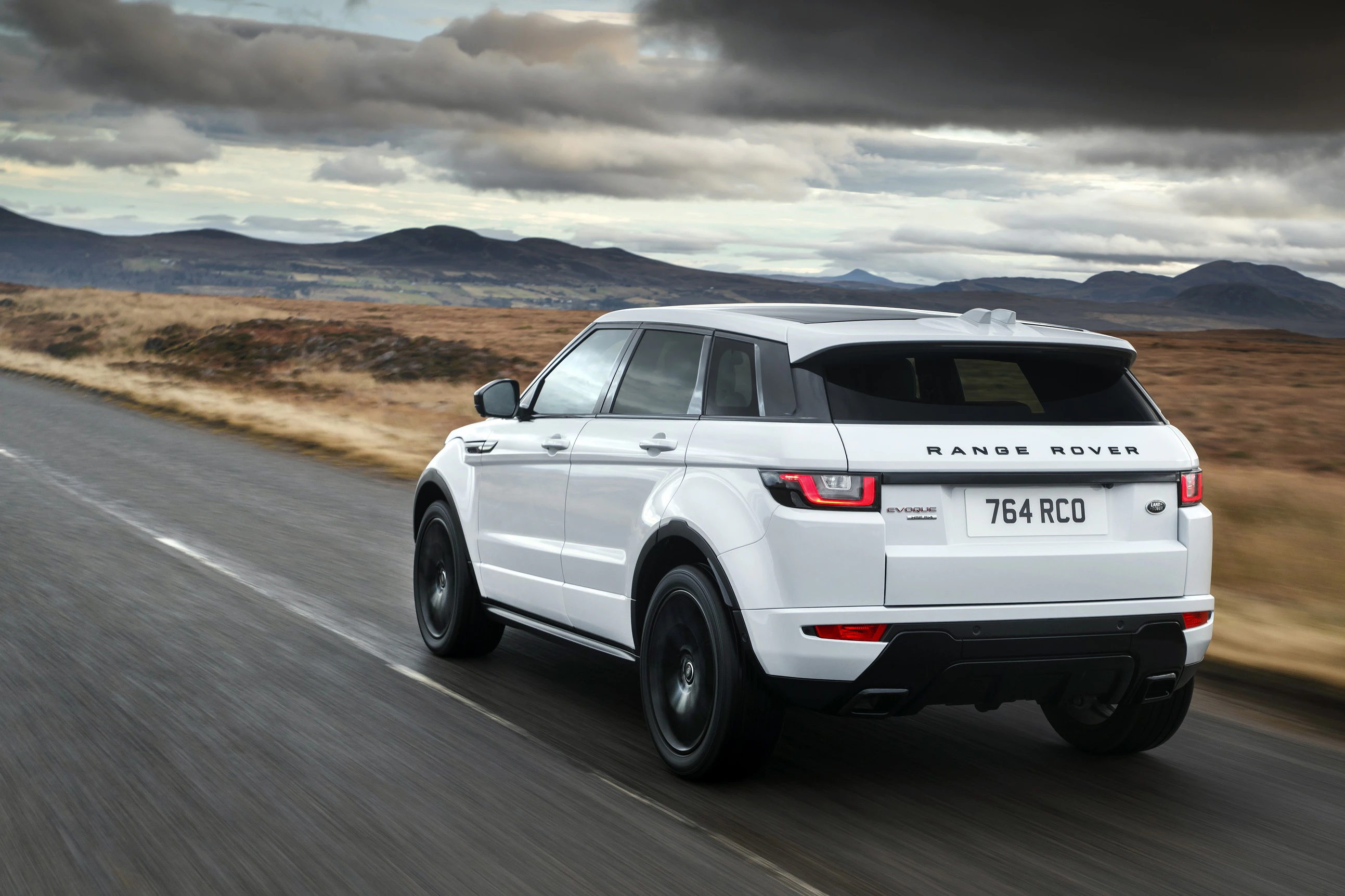 Weißer Range Rover Evoque auf Landstraße, fährt durch eine offene Landschaft mit Bergen im Hintergrund unter bewölktem Himmel.