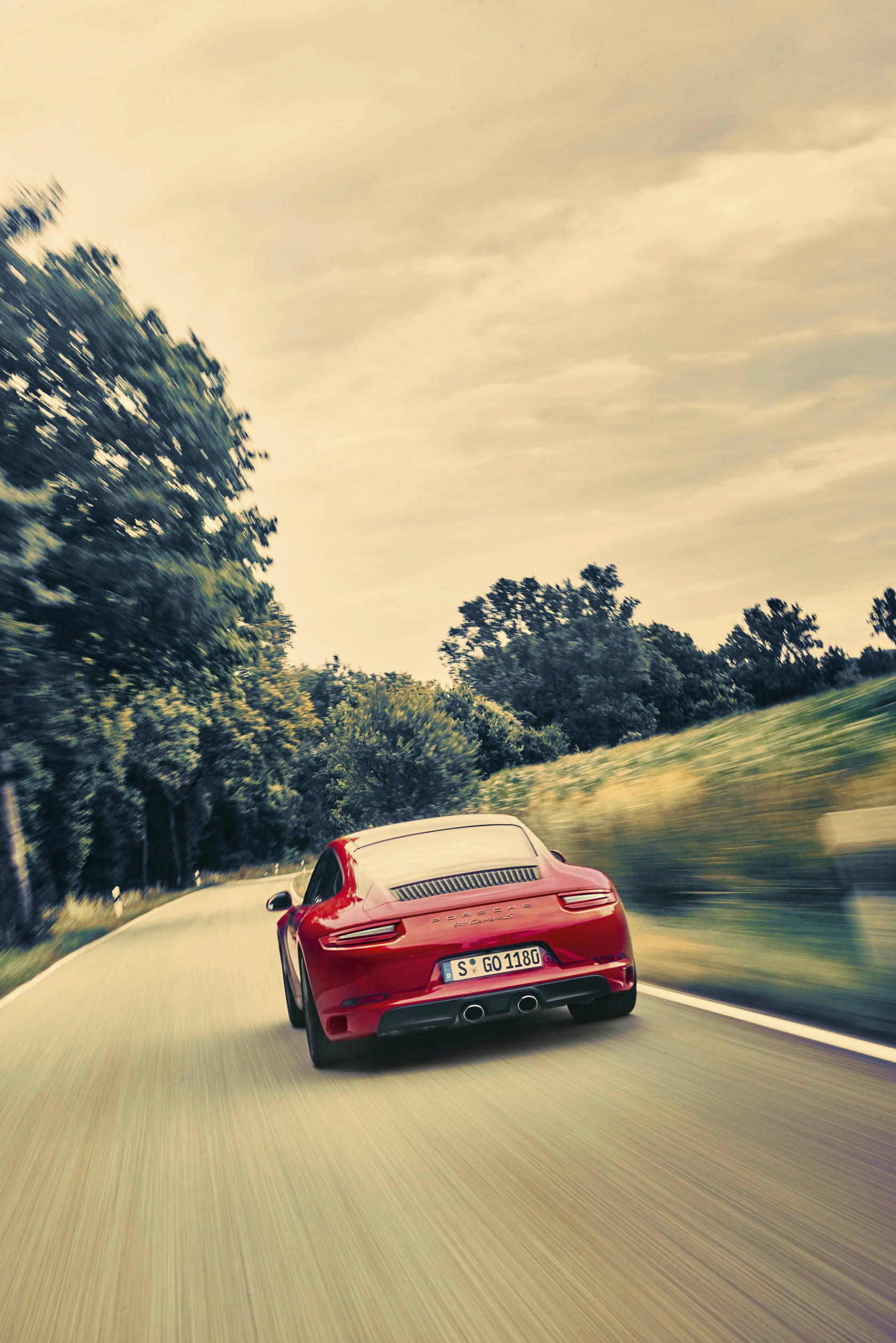 Roter Sportwagen fährt über eine landschaftliche Straße, umgeben von Bäumen und bewölktem Himmel.