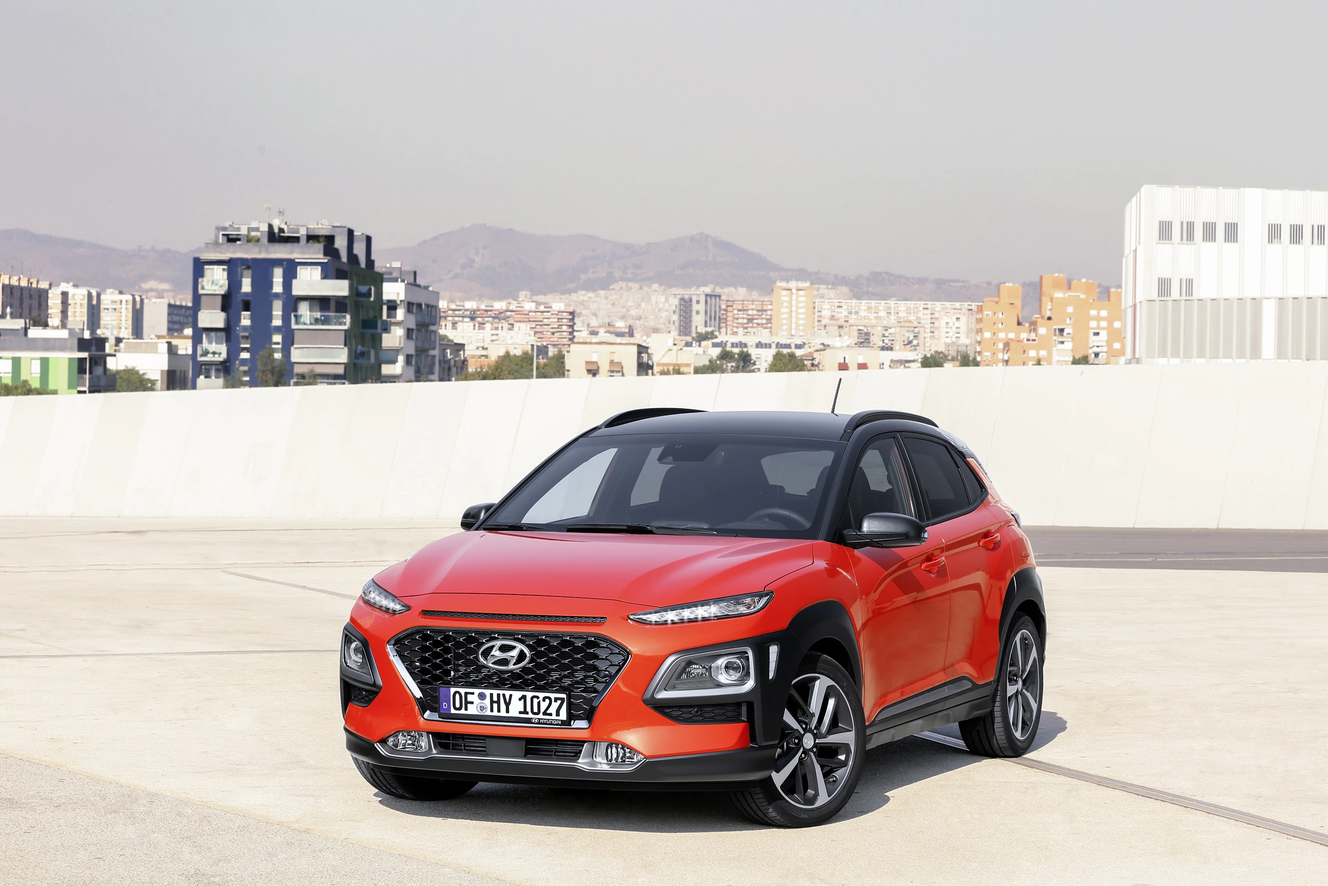 Ein rotes Auto, Hyundai Kona, steht auf einem Parkplatz mit städtischem Hintergrund und bewölktem Himmel.