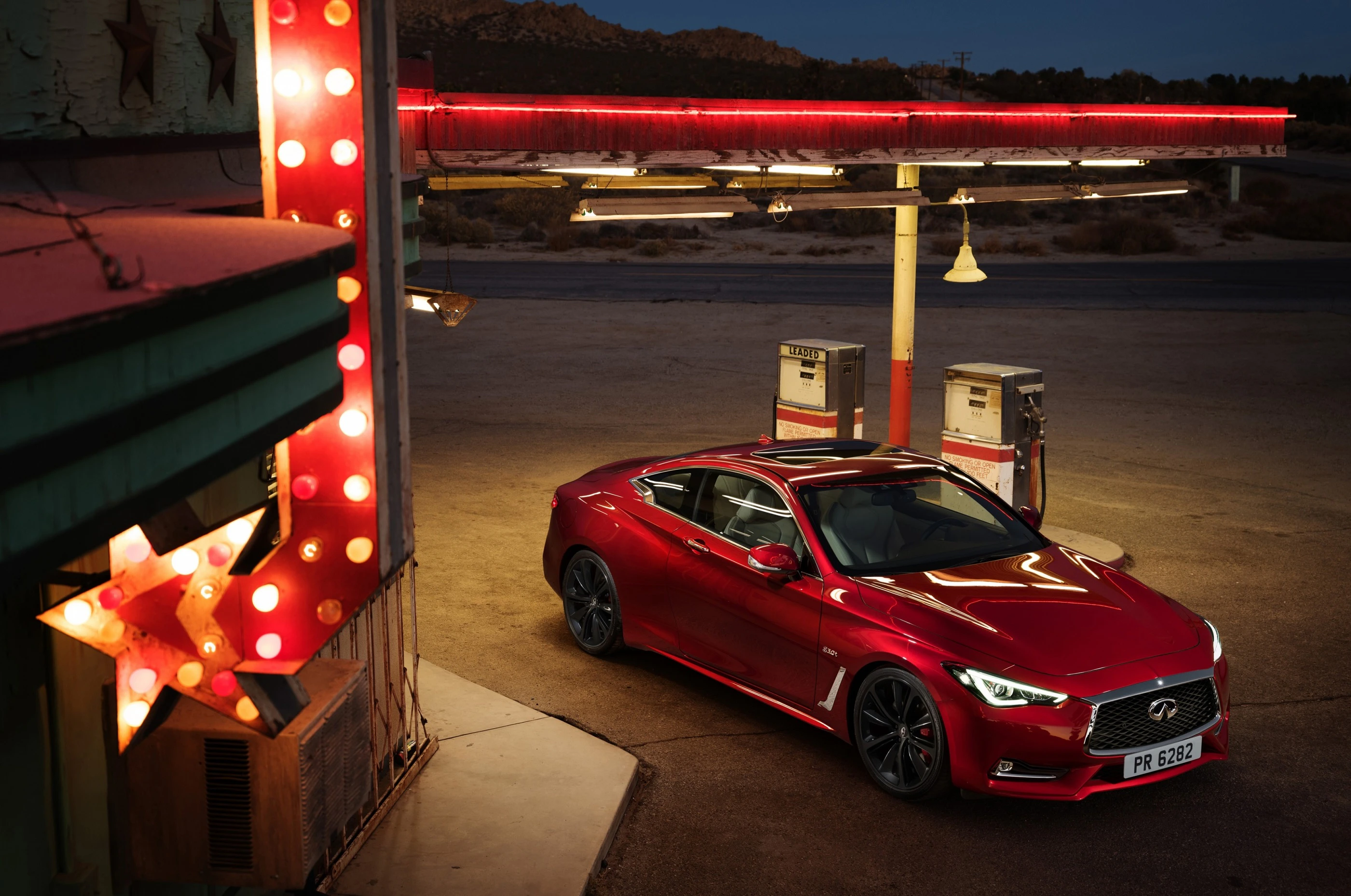 Rotes Auto an alter Tankstelle bei Dämmerung, umgeben von leuchtenden Schildern und rustikalem Ambiente.