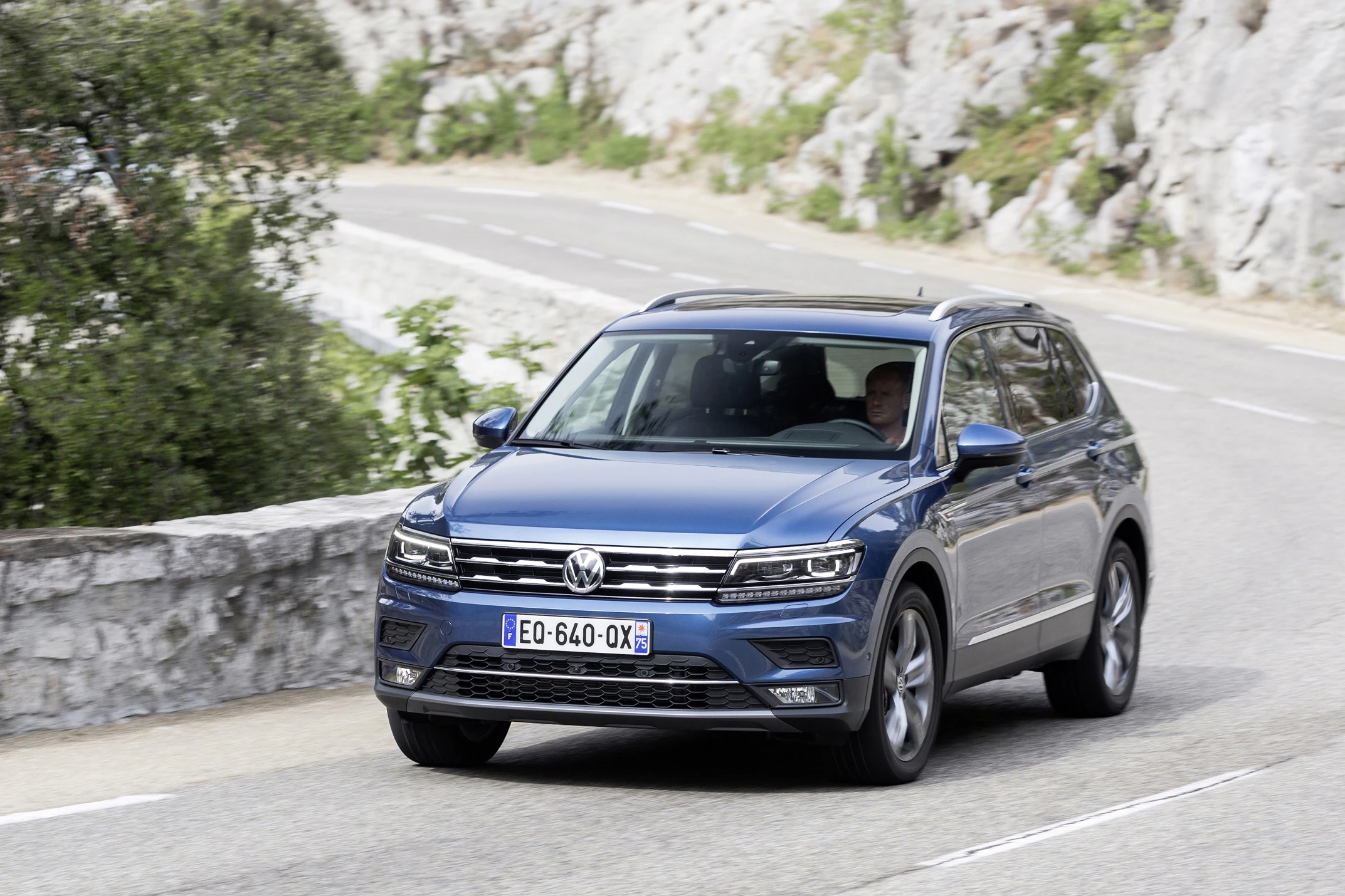 Blauer VW Tiguan fährt auf einer kurvenreichen, bergigen Straße. Fahrer am Steuer, umgeben von Felsen und Bäumen.