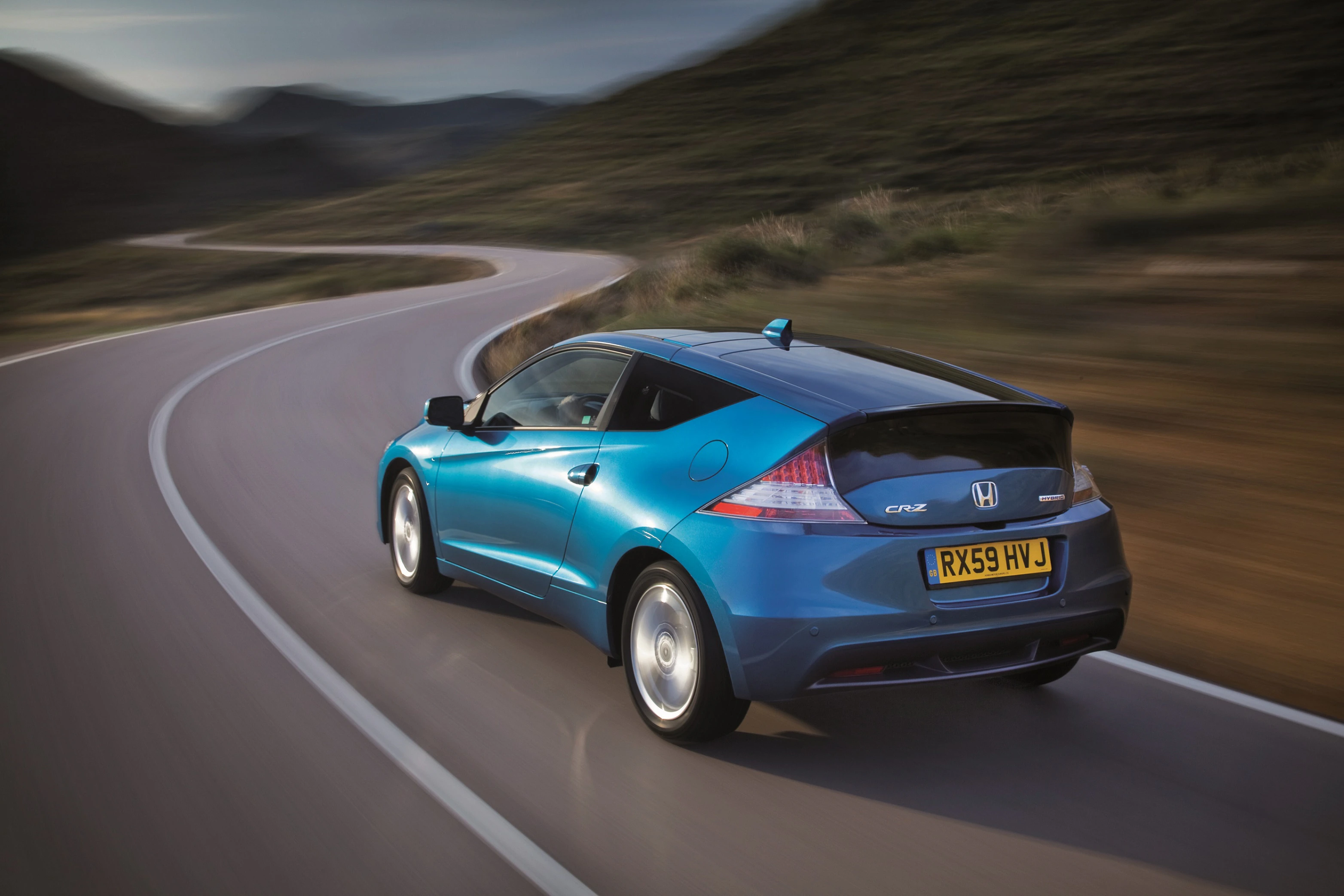 Blauer Honda CR-Z fährt auf kurviger Straße in ländlicher Landschaft, im Sonnenlicht. Dynamische Bewegung und sportliches Design.