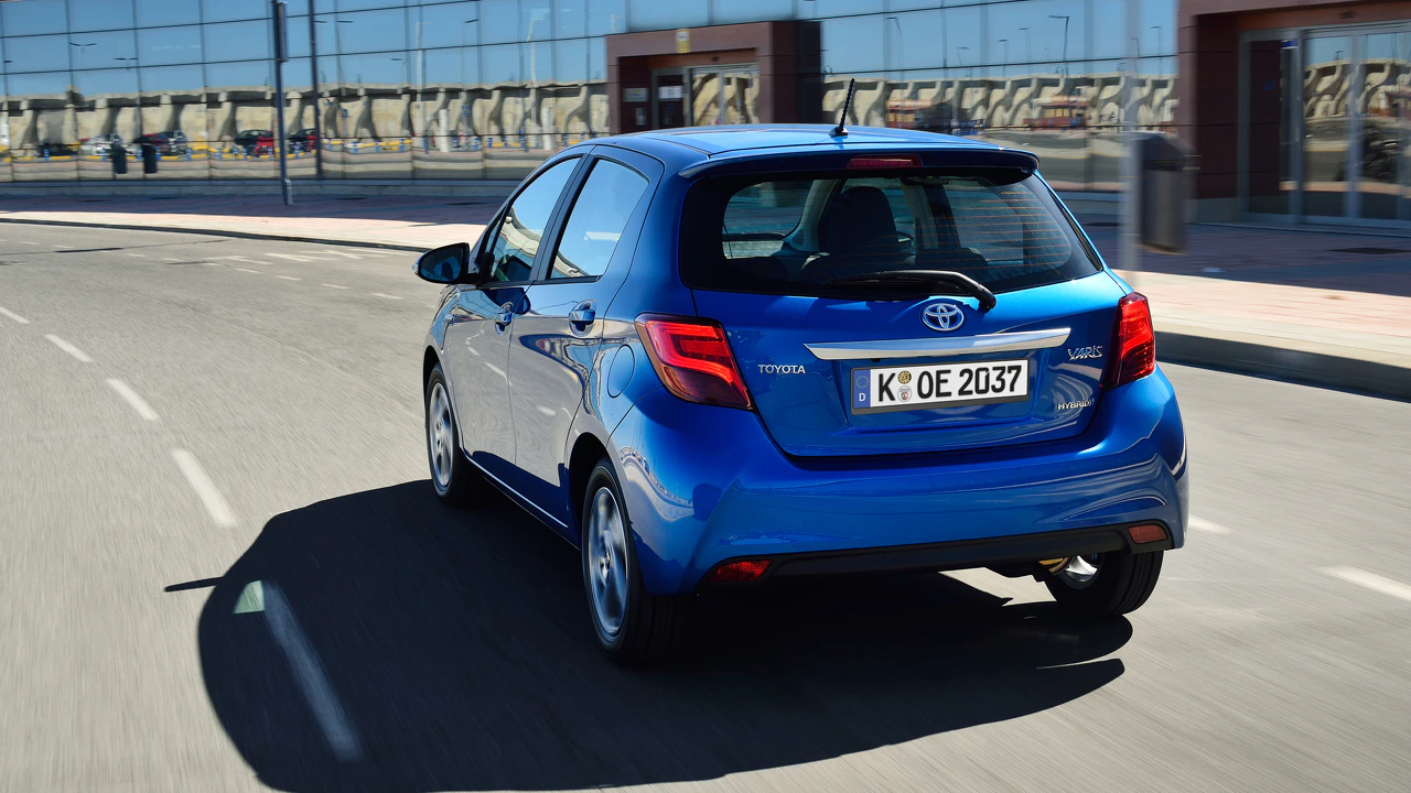 Ein blauer Toyota Yaris fährt auf einer Straße. Im Hintergrund spiegelt ein modernes Gebäude den Himmel. Klares Wetter, tagsüber.