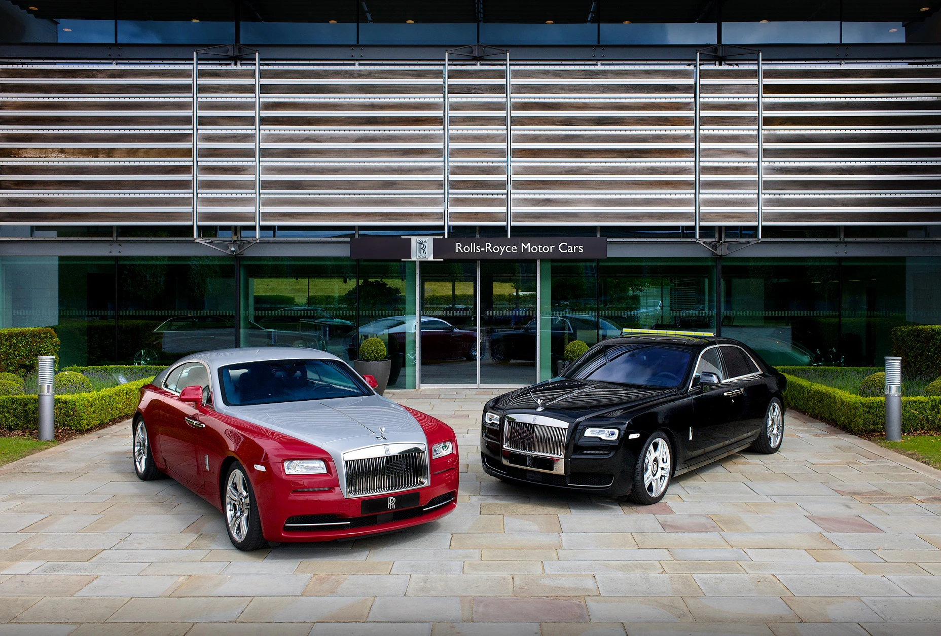 Zwei Rolls-Royce-Autos vor einem modernen Gebäude: links ein rotes Coupé und rechts eine schwarze Limousine.