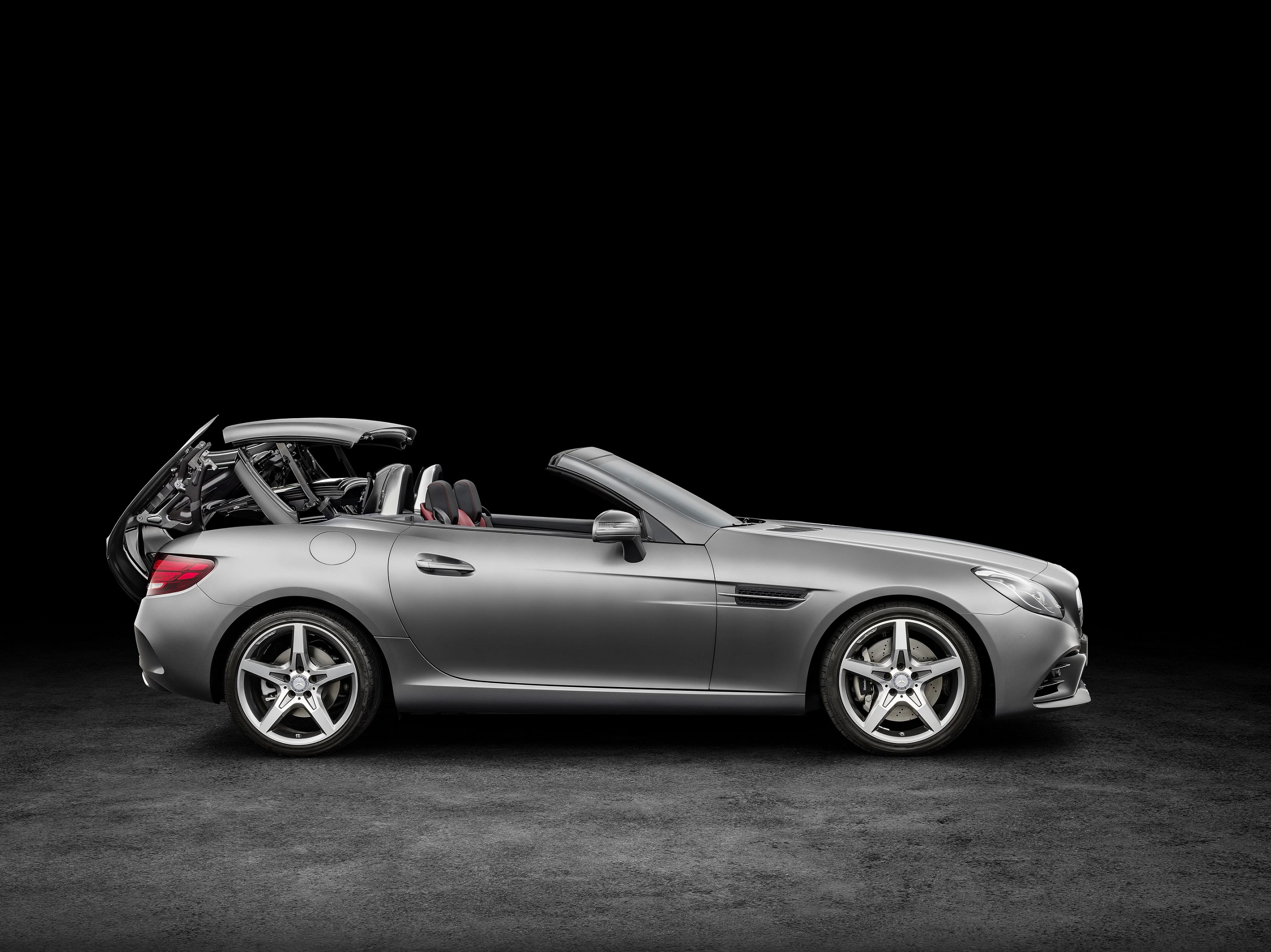 Silbernes Cabrio mit offenem Verdeck in Seitenansicht vor schwarzem Hintergrund, zeigt sportliches Design und moderne Felgen.