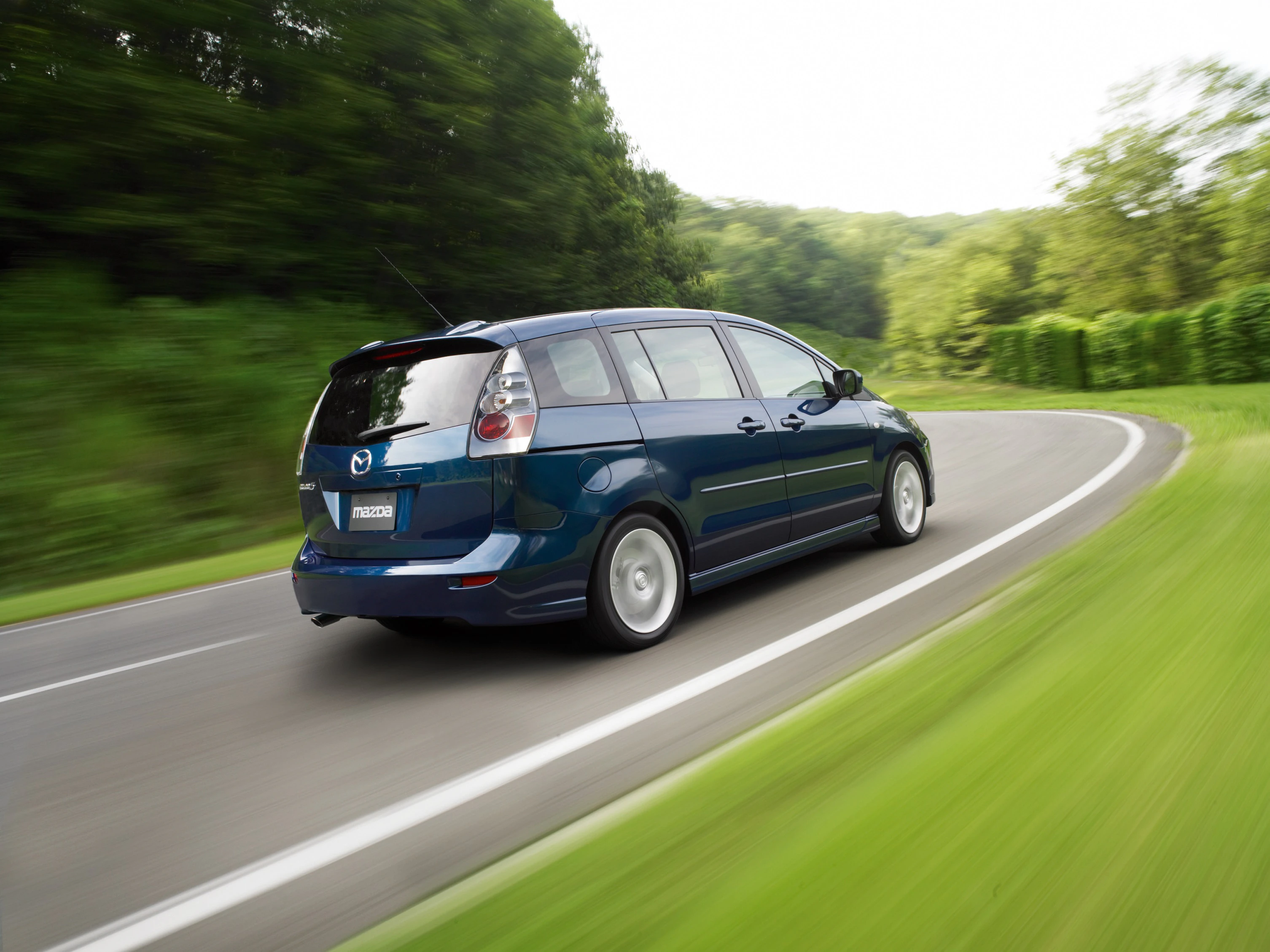 Blauer Minivan auf einer kurvigen Landstraße, umgeben von grüner Landschaft und Bäumen, fährt schnell nach rechts.