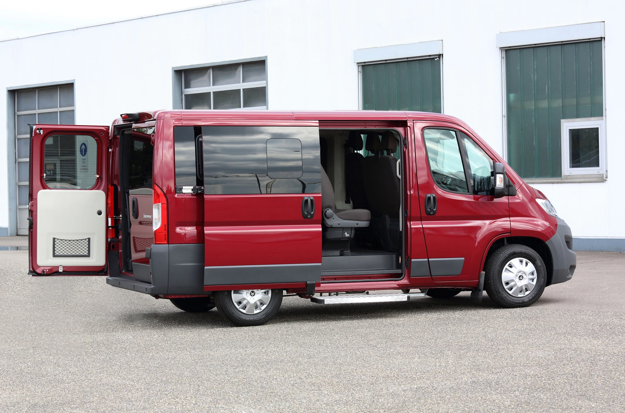 Roter Lieferwagen mit geöffneten Türen, seitlich auf einem Parkplatz vor einem Gebäude stehend.
