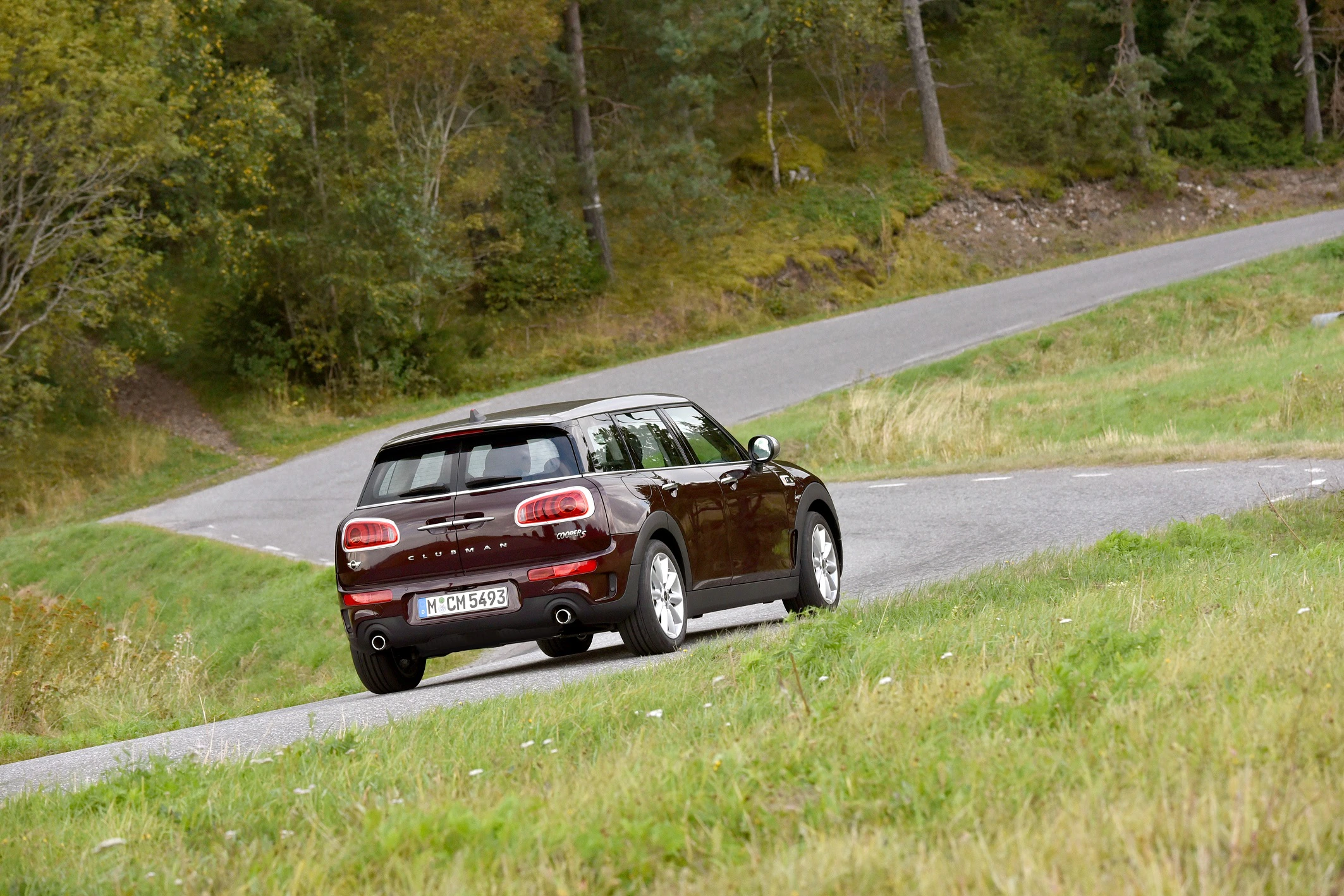 Ein roter Mini Cooper Clubman fährt auf einer gewundenen Landstraße durch eine grüne, bewaldete Landschaft.