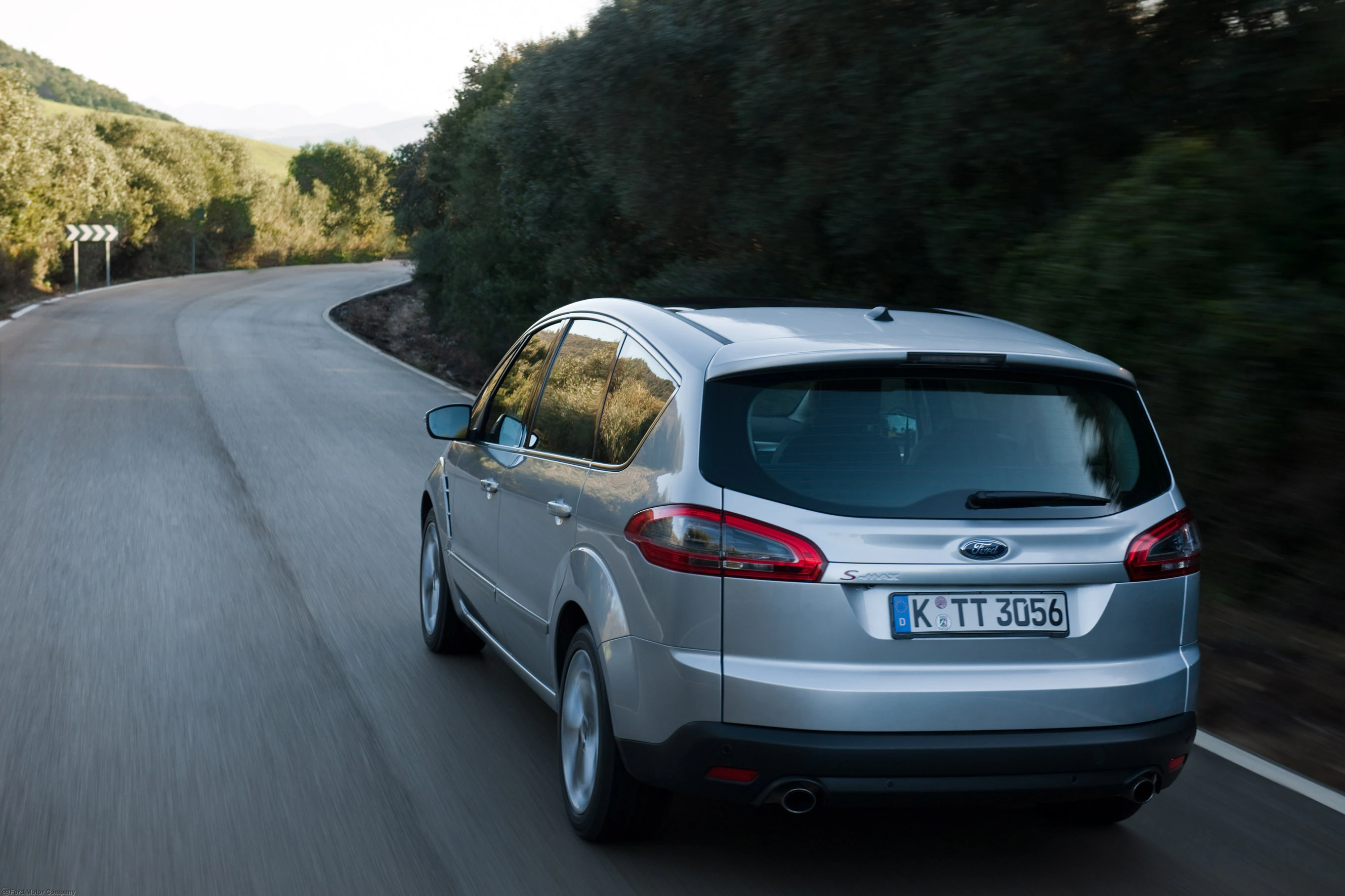 Silberner Ford S-MAX fährt auf einer kurvigen Landstraße durch eine grüne Landschaft. Fahrzeugkennzeichen: K-TT 3056.