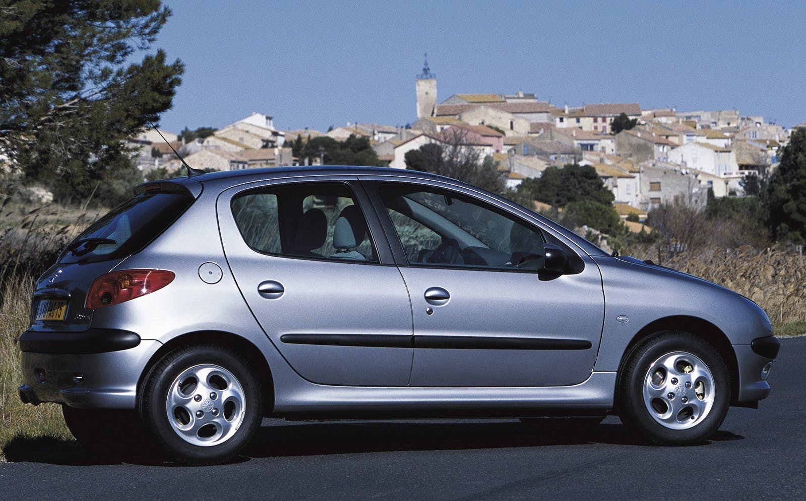 Silberner Kleinwagen steht auf einer Straße vor einem Dorf mit Hügeln im Hintergrund an einem klaren, sonnigen Tag.