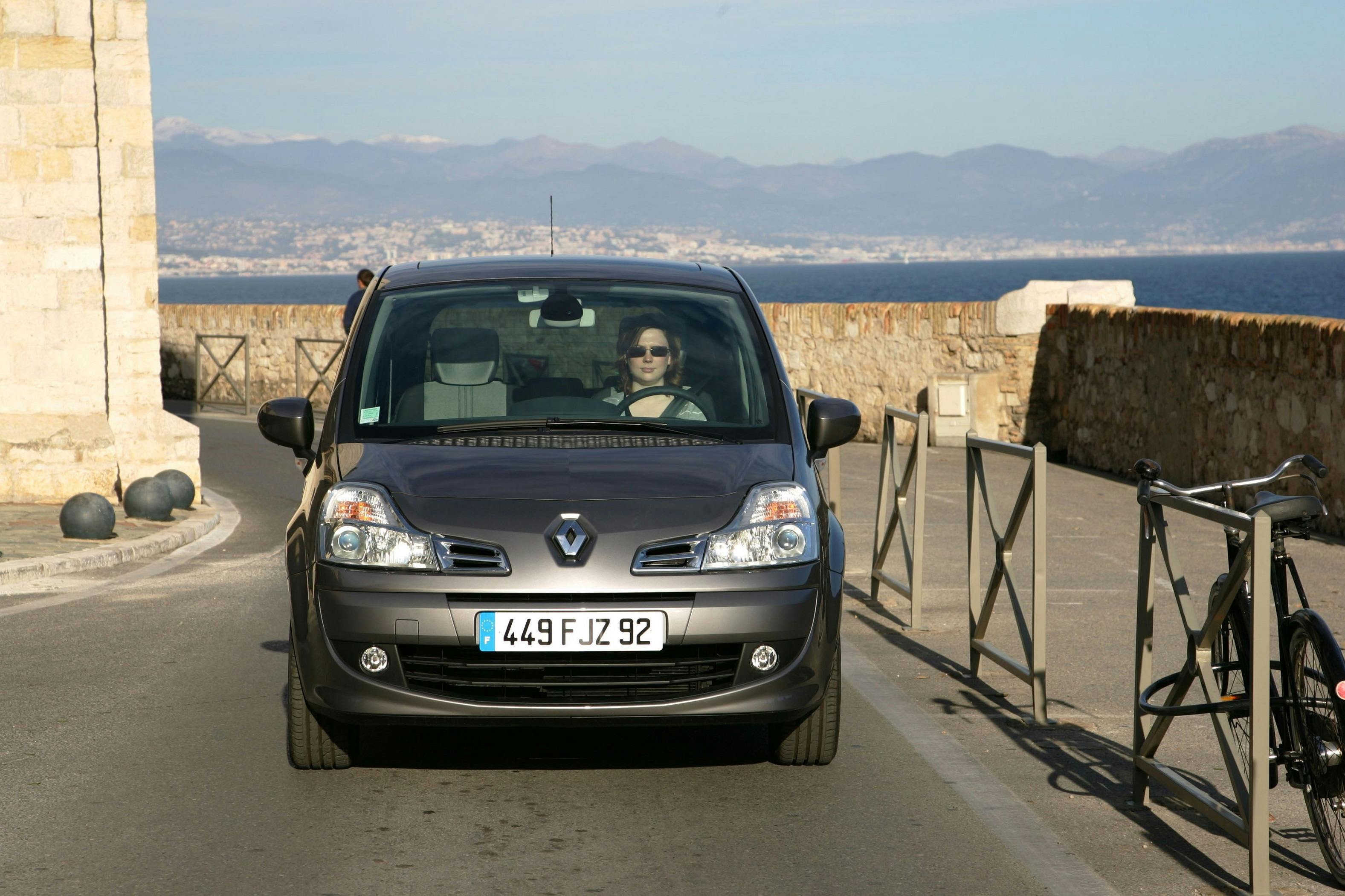 Auto fährt auf Küstenstraße; Fahrer trägt Sonnenbrille. Im Hintergrund Meer und Berglandschaft. Fahrrad an der Seite.