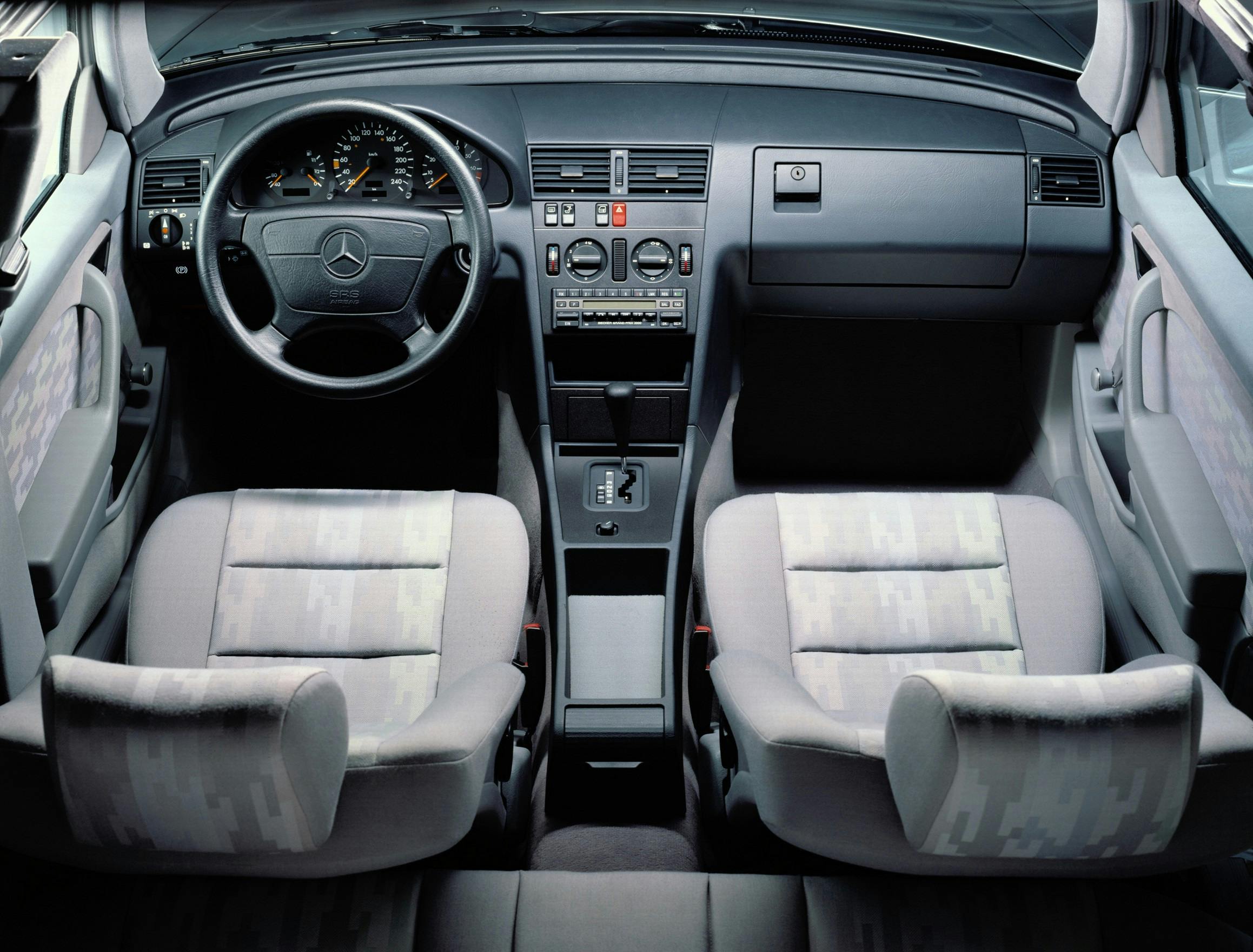 Innenraum eines Autos mit Mercedes-Benz-Logo, grauen Stoffpolstern, Lenkrad links, Armaturenbrett mit Radio und Klimaanlage.