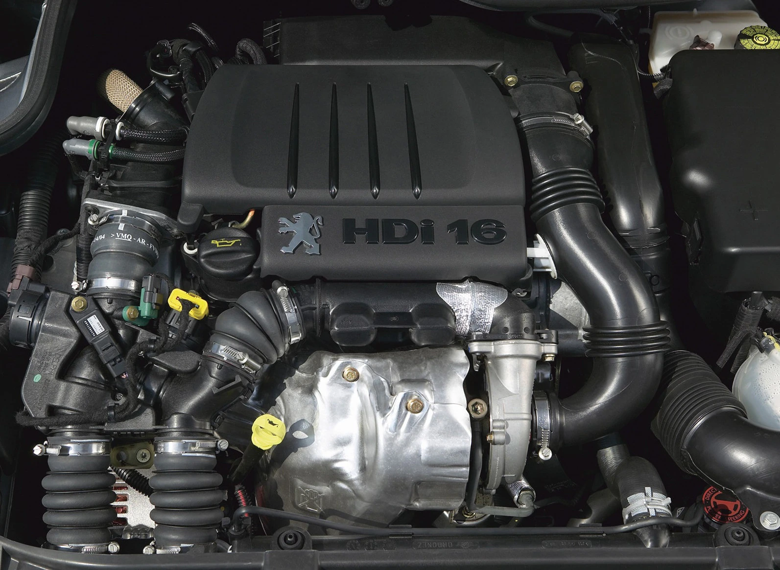 Nahaufnahme eines HDi 16 Dieselmotors in einem Auto mit verschiedenen sichtbaren Komponenten und dem Peugeot-Logo auf der Abdeckung.