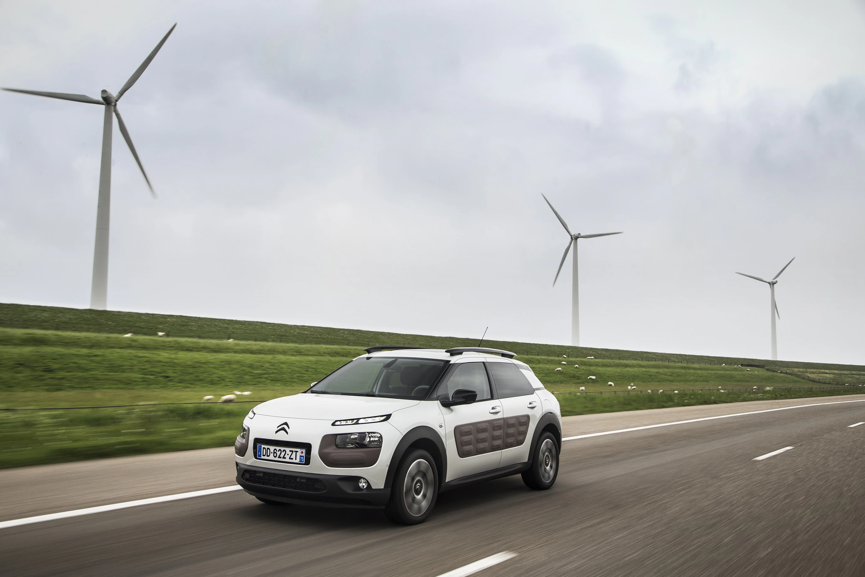 Auto fährt auf Landstraße, umgeben von Windturbinen auf grünen Feldern unter bewölktem Himmel.