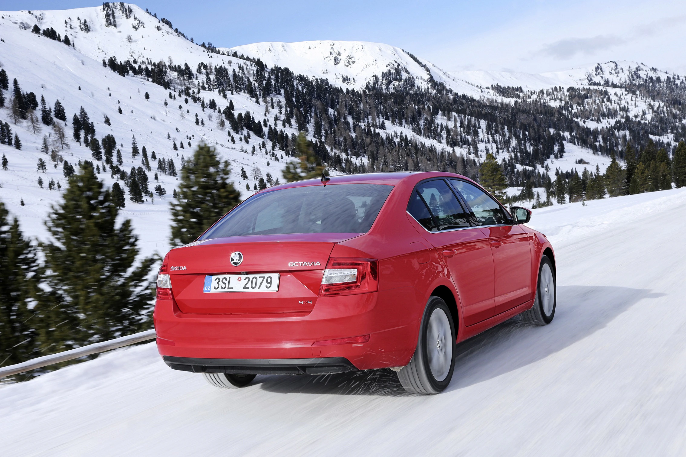 Rotes Auto fährt auf verschneiter Bergstraße, umgeben von Tannen und schneebedeckten Bergen, unter bewölktem Himmel.