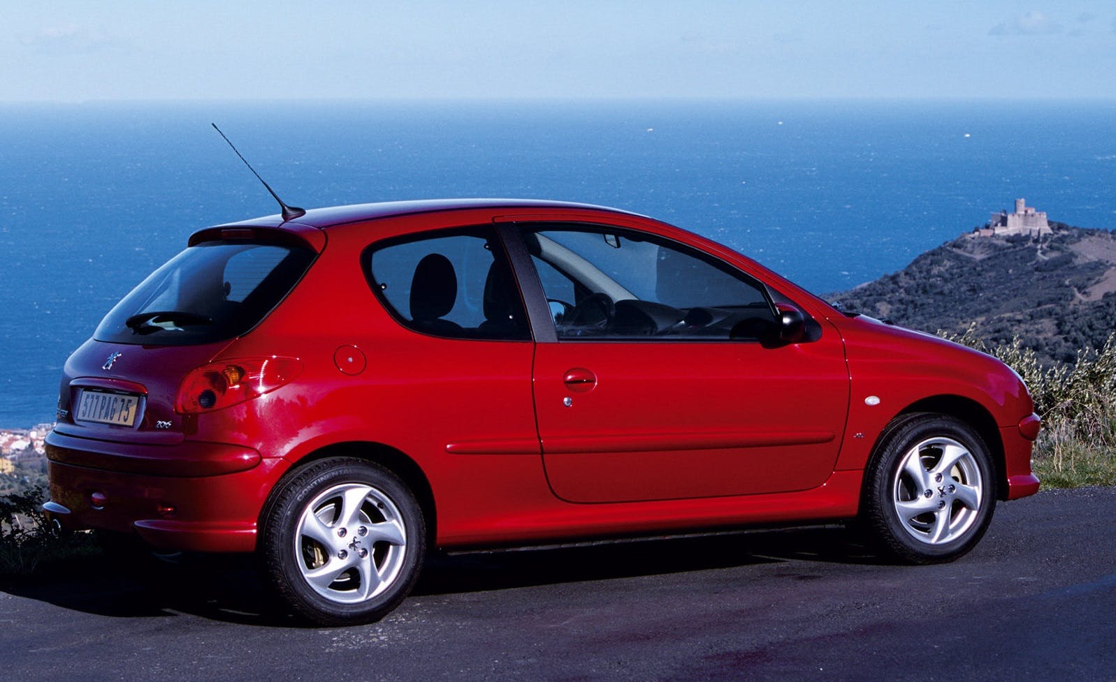 Rotes Auto geparkt vor einer Küstenlandschaft mit blauem Meer und einem Hügel im Hintergrund, auf dem eine kleine Burg steht.