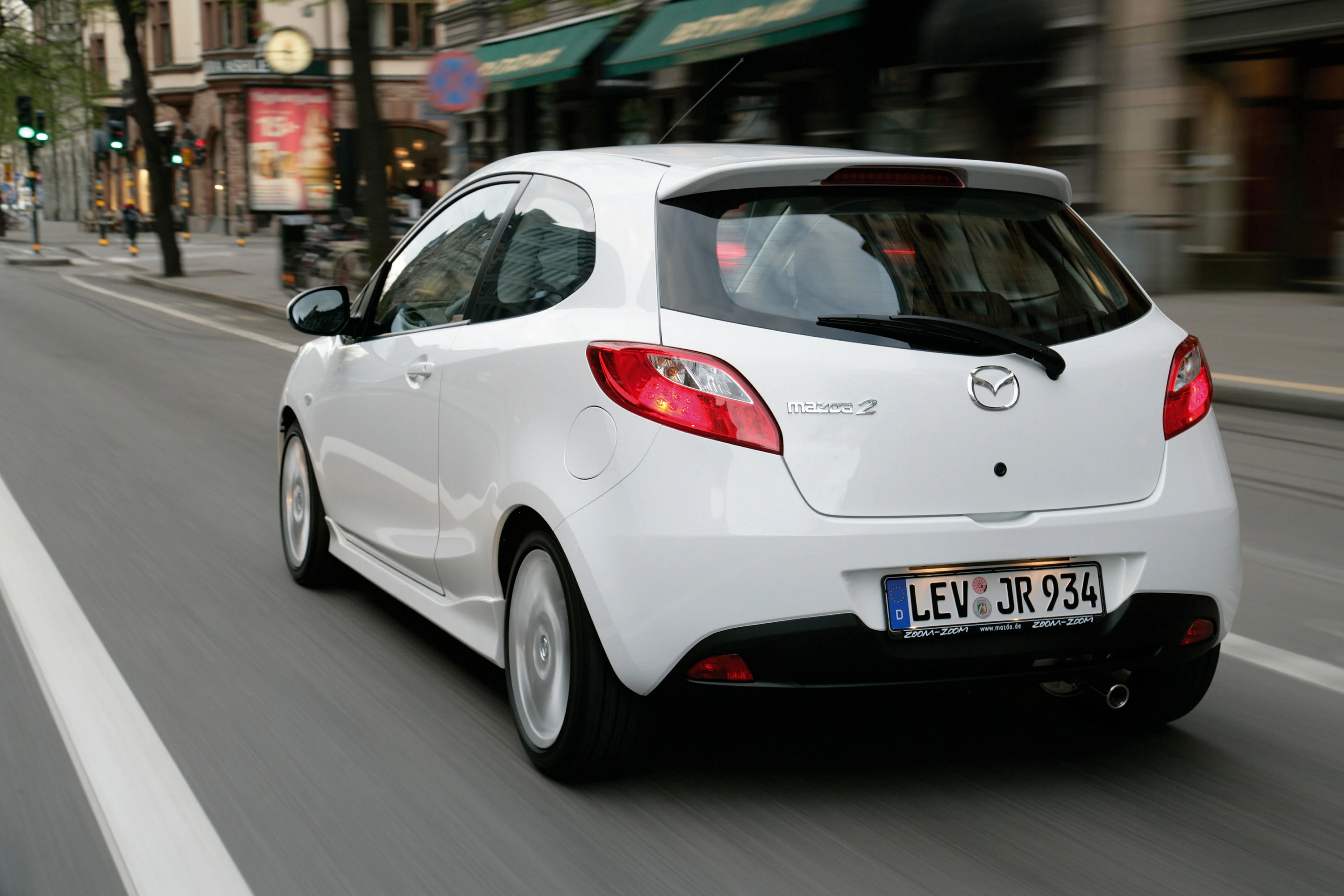 Weißer Mazda 2 fährt auf einer Stadtstraße, umgeben von historischen Gebäuden. Das Auto ist von hinten zu sehen, mit der Nummernschild "LEV JR 934".