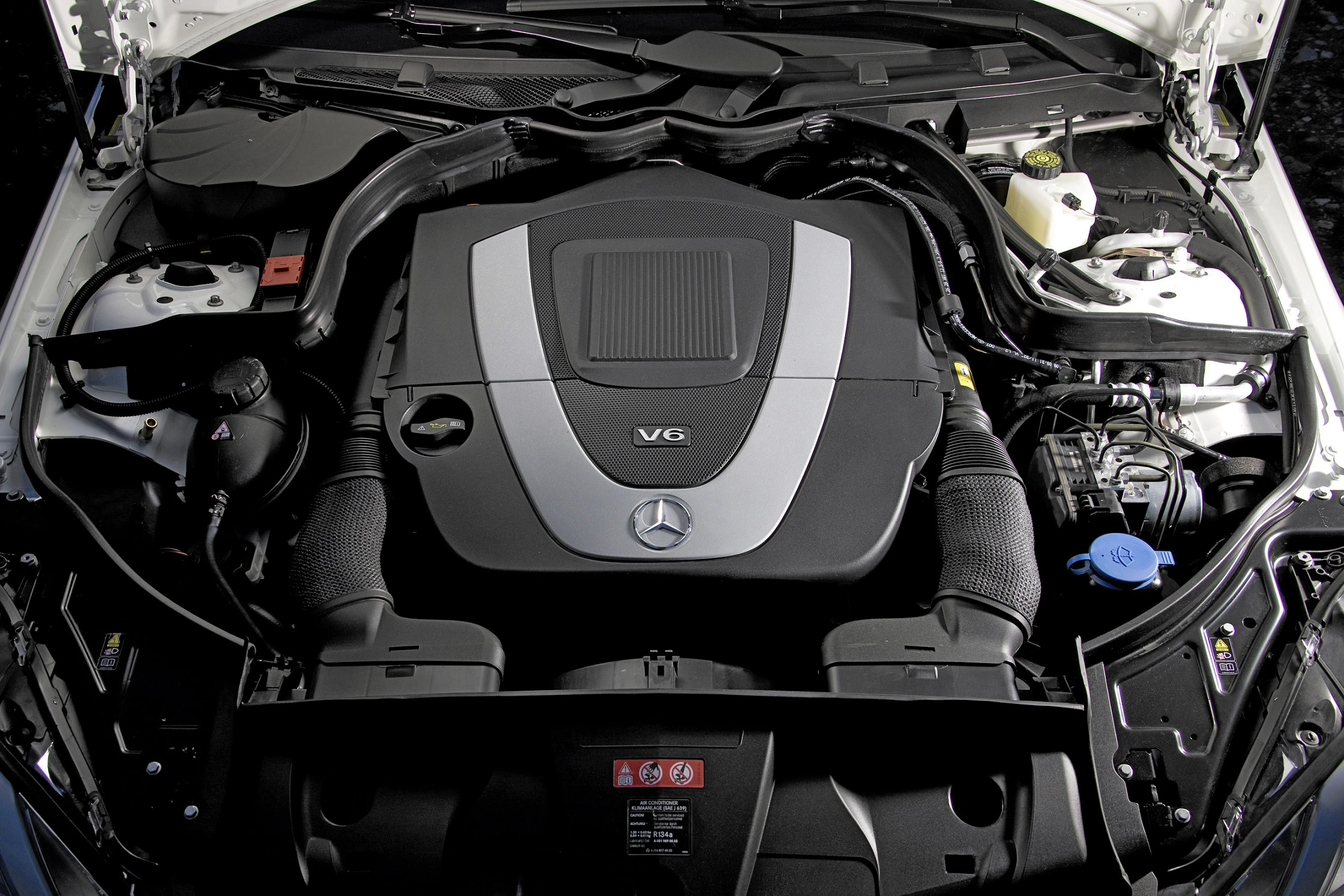 Motorraum eines Mercedes-Fahrzeugs mit V6-Motor. Sichtbar sind Motorabdeckung, Kühlmittelbehälter und andere technische Komponenten.