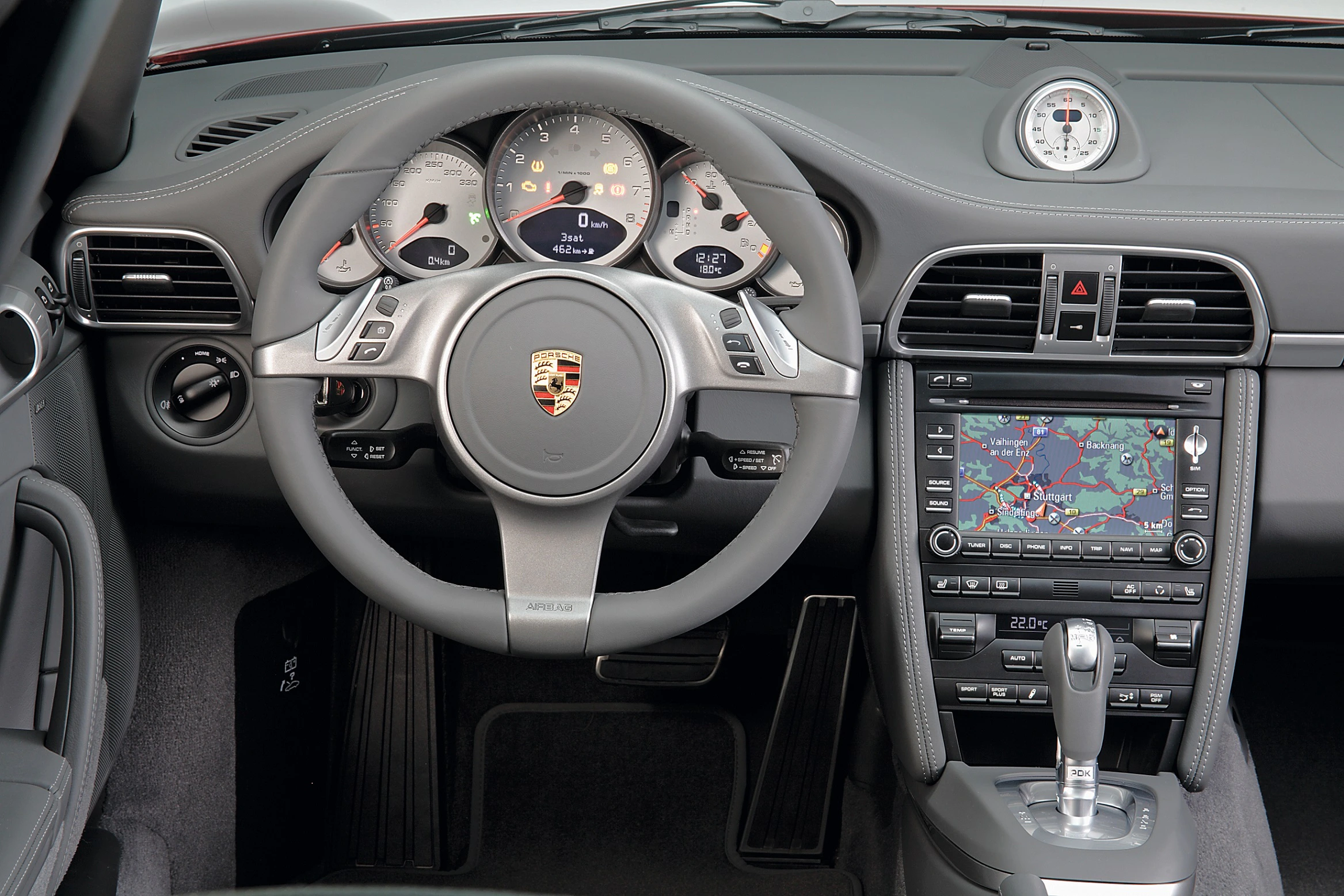 Innenraum eines Sportwagens mit Porsche-Logo auf dem Lenkrad, Tachometer, Navigationsanzeige und zentralem Schalthebel.