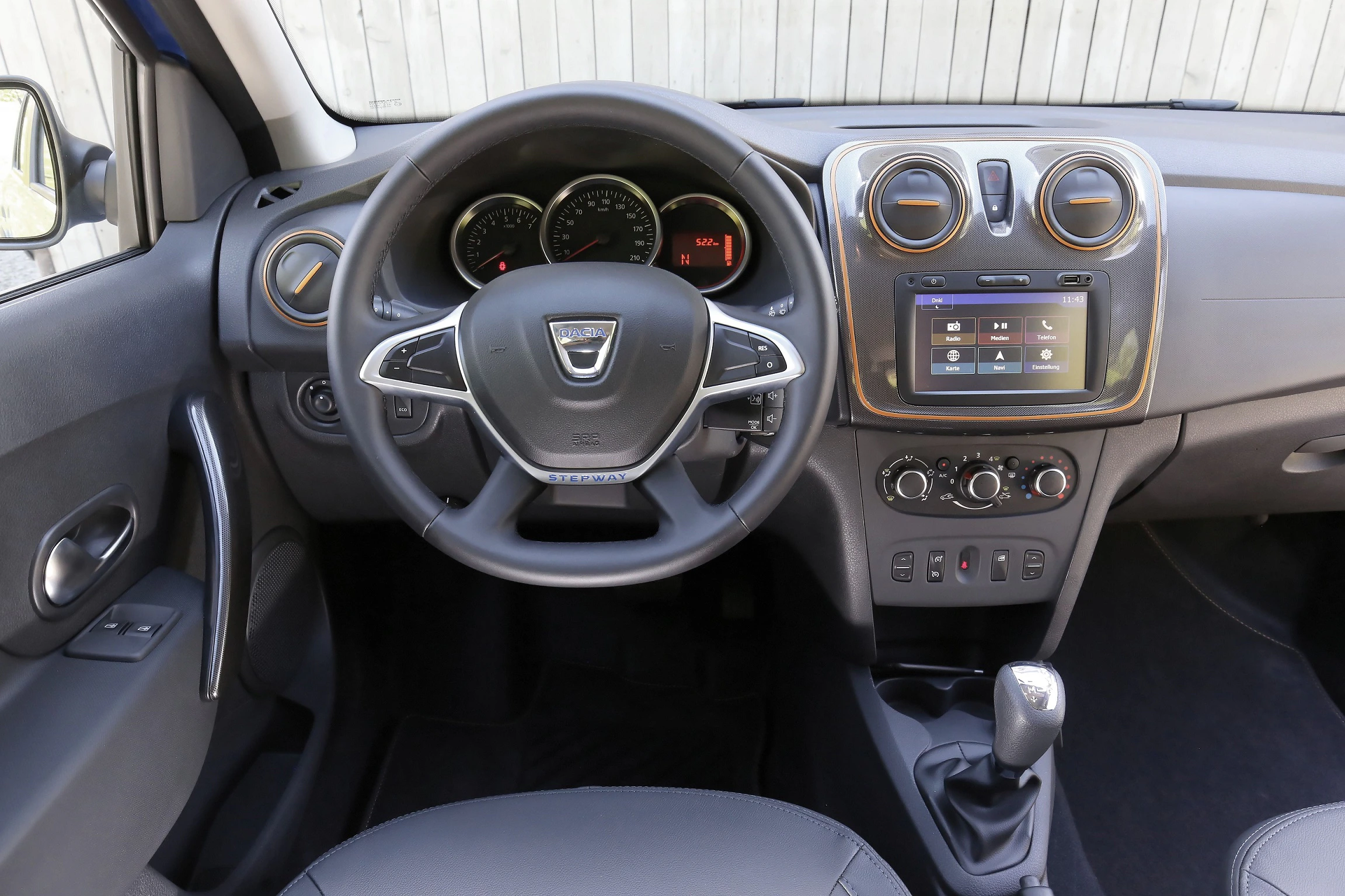 Innenansicht eines Autos mit Lenkrad, Armaturenbrett, Tachometer und Multimedia-Display. Moderne Steuerungselemente sichtbar.