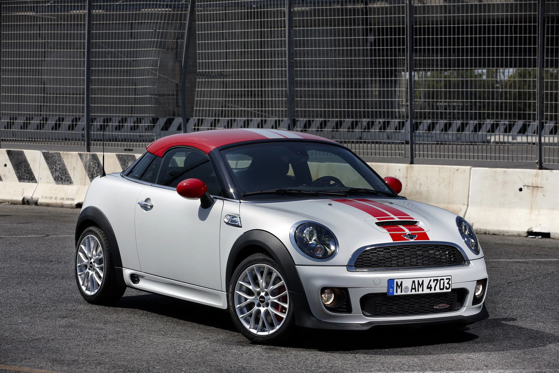 Silberner Sportwagen mit roten Akzenten und Streifen, Mini Cooper Modell, parkt vor einer hohen Metallzaunwand auf der Straße.