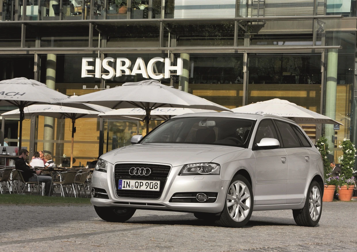 Silberner Audi A3 geparkt vor einem Café mit großen Sonnenschirmen, Aufschrift "Eisbach" sichtbar im Hintergrund.