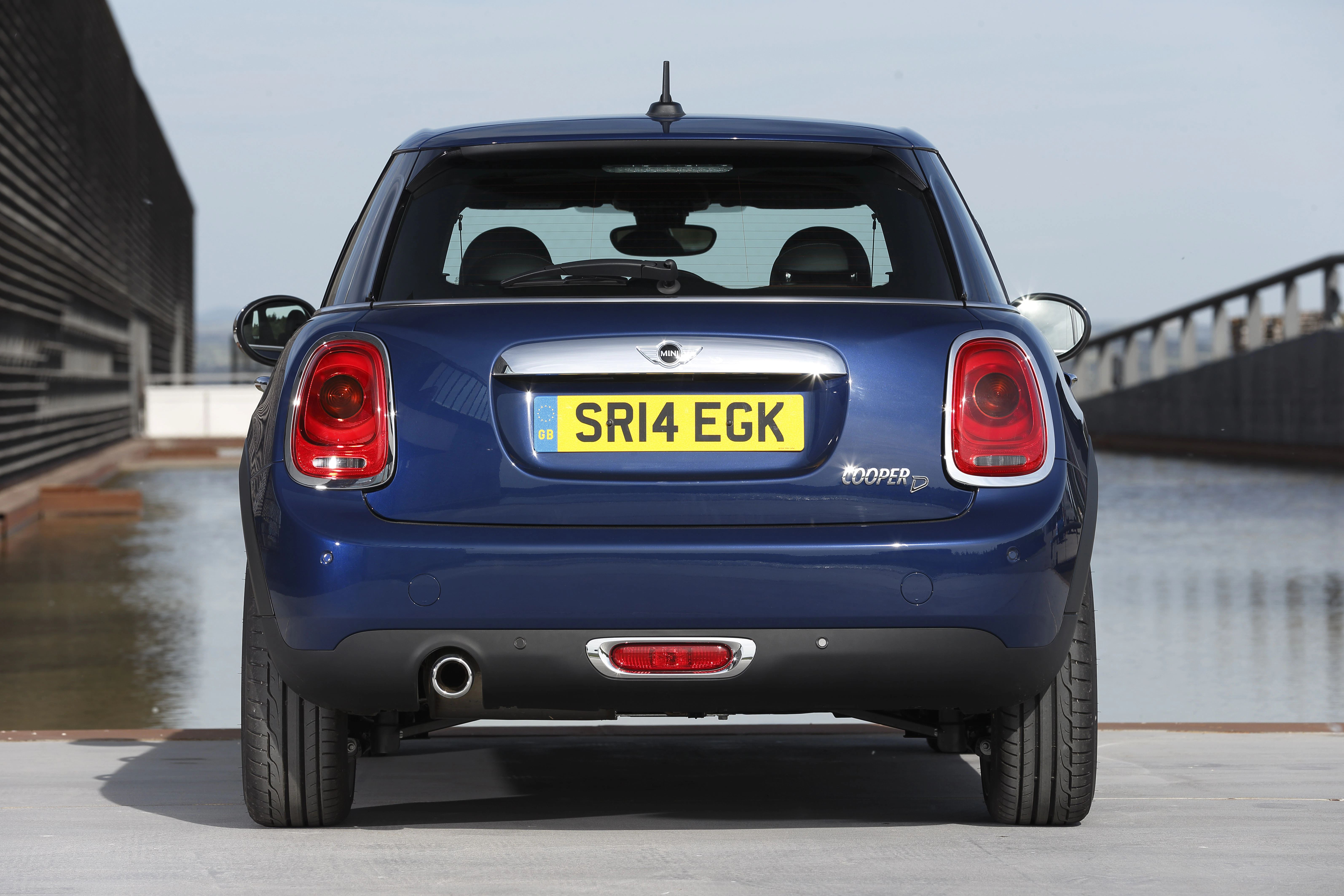 Blauer Mini Cooper mit Nummernschild SR14 EGK, Heckansicht auf einer Brücke mit Wasser im Hintergrund.