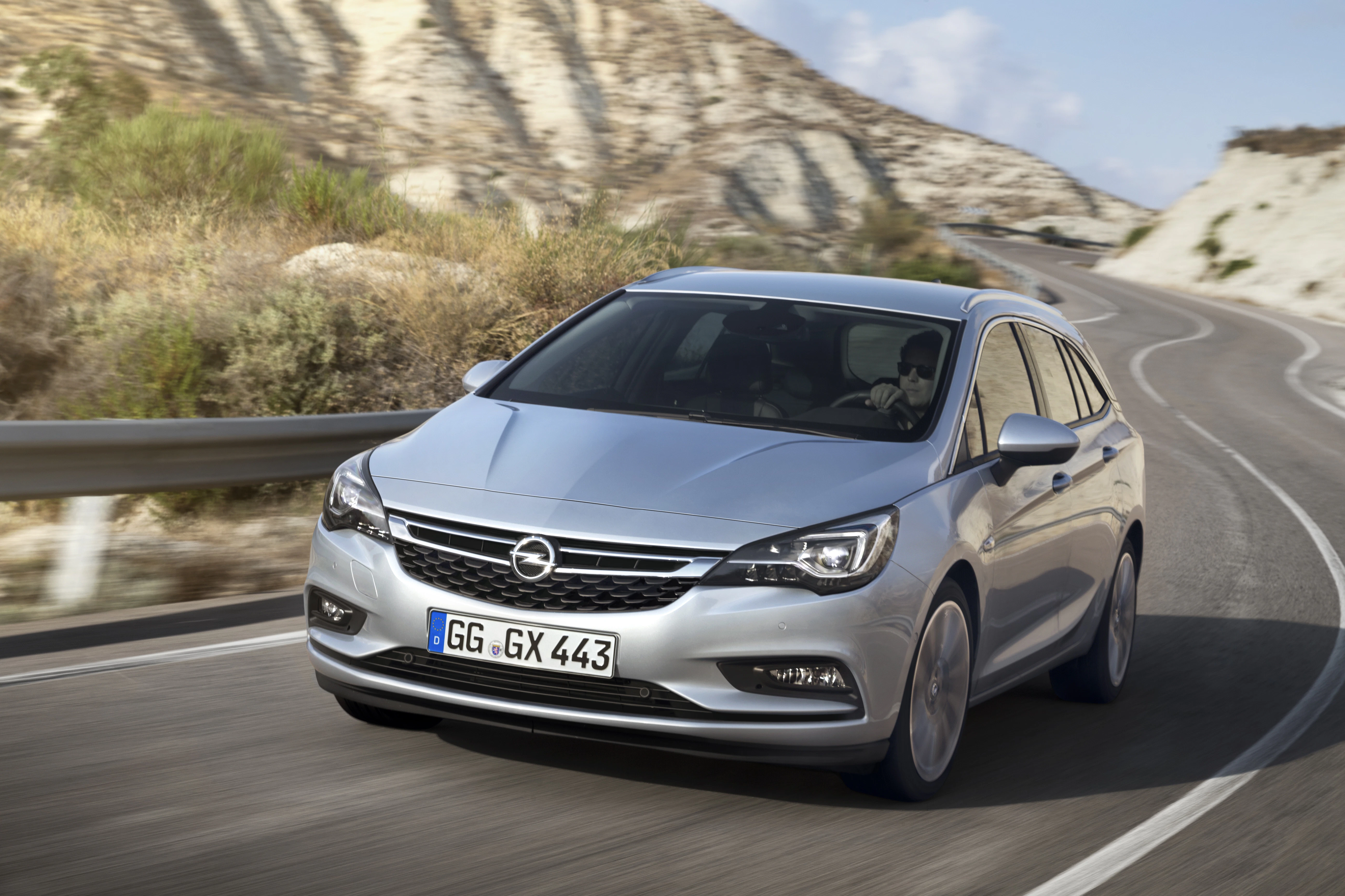 Silberner Opel Astra fährt auf gewundener Straße in einer trockenen, bergigen Landschaft.