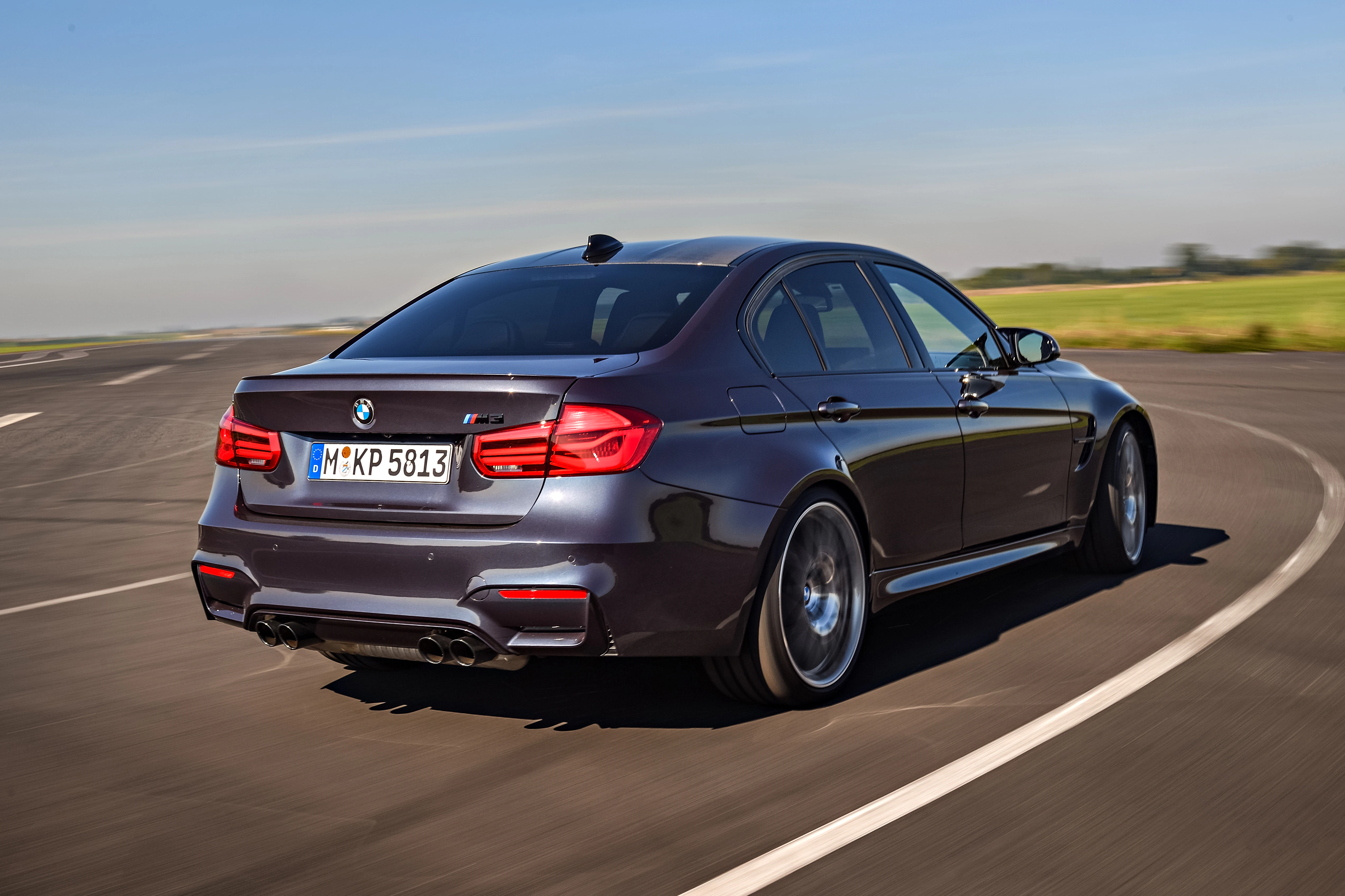 Grauer BMW M3 fährt auf kurviger Landstraße bei sonnigem Wetter, grüne Felder im Hintergrund.