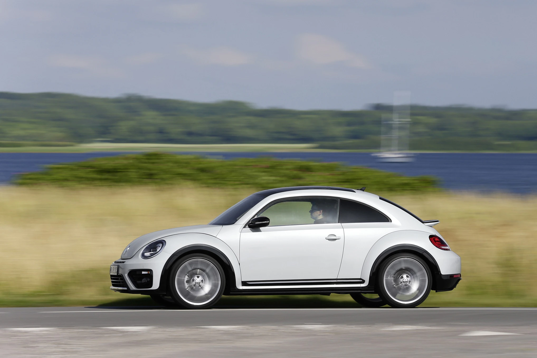 Weißer VW Beetle fährt auf einer Landstraße, im Hintergrund grüne Felder und ein See.