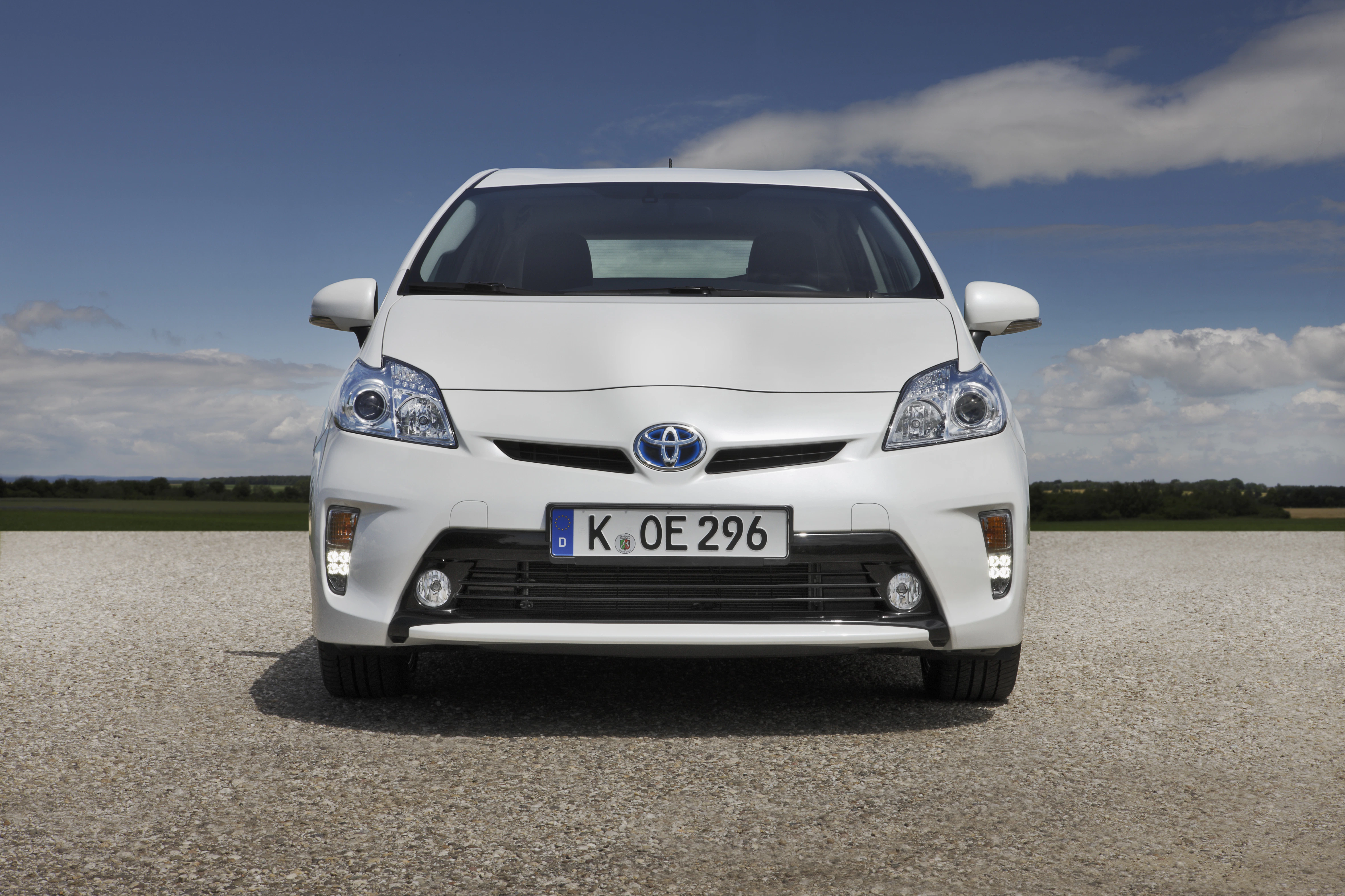 Weißer Toyota Prius von vorne, geparkt auf einem Kiesweg. Blaue Himmel und Wolken im Hintergrund.