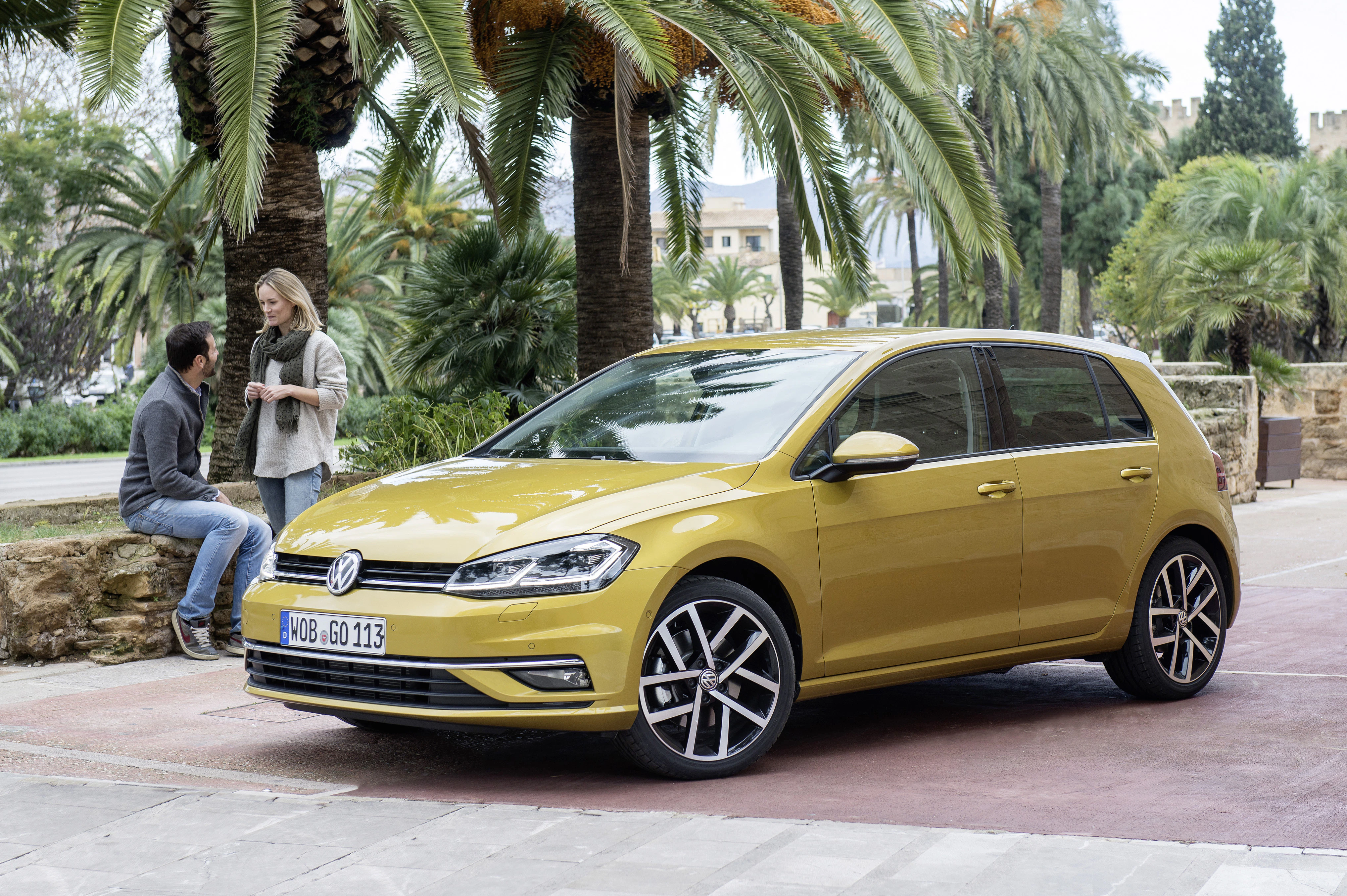 Ein gelber VW Golf steht vor Palmen; im Hintergrund unterhalten sich zwei Personen.