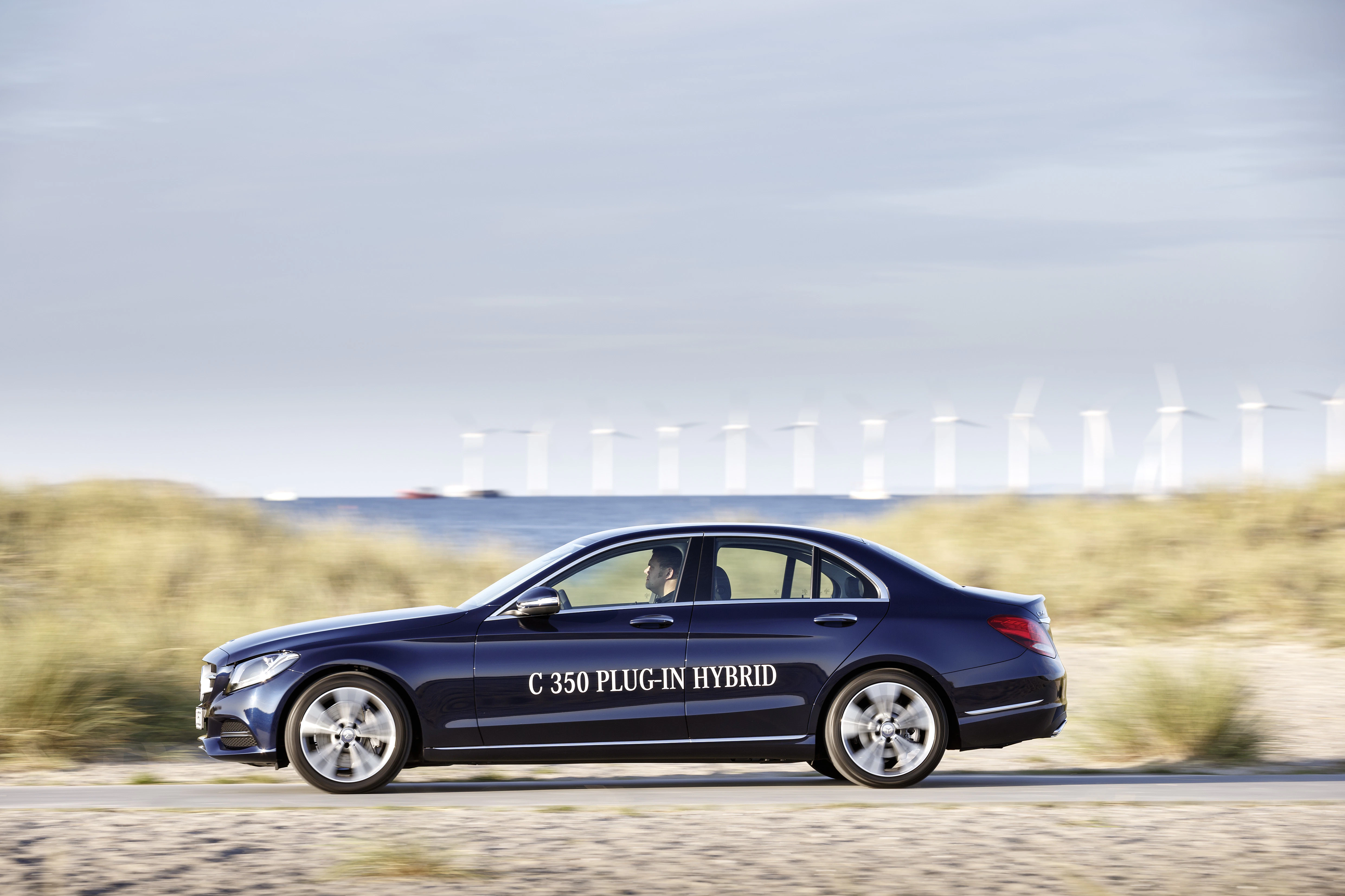Blaues Auto mit der Aufschrift "C 350 PLUG-IN HYBRID" fährt an einer Küstenlandschaft mit Windrädern vorbei.