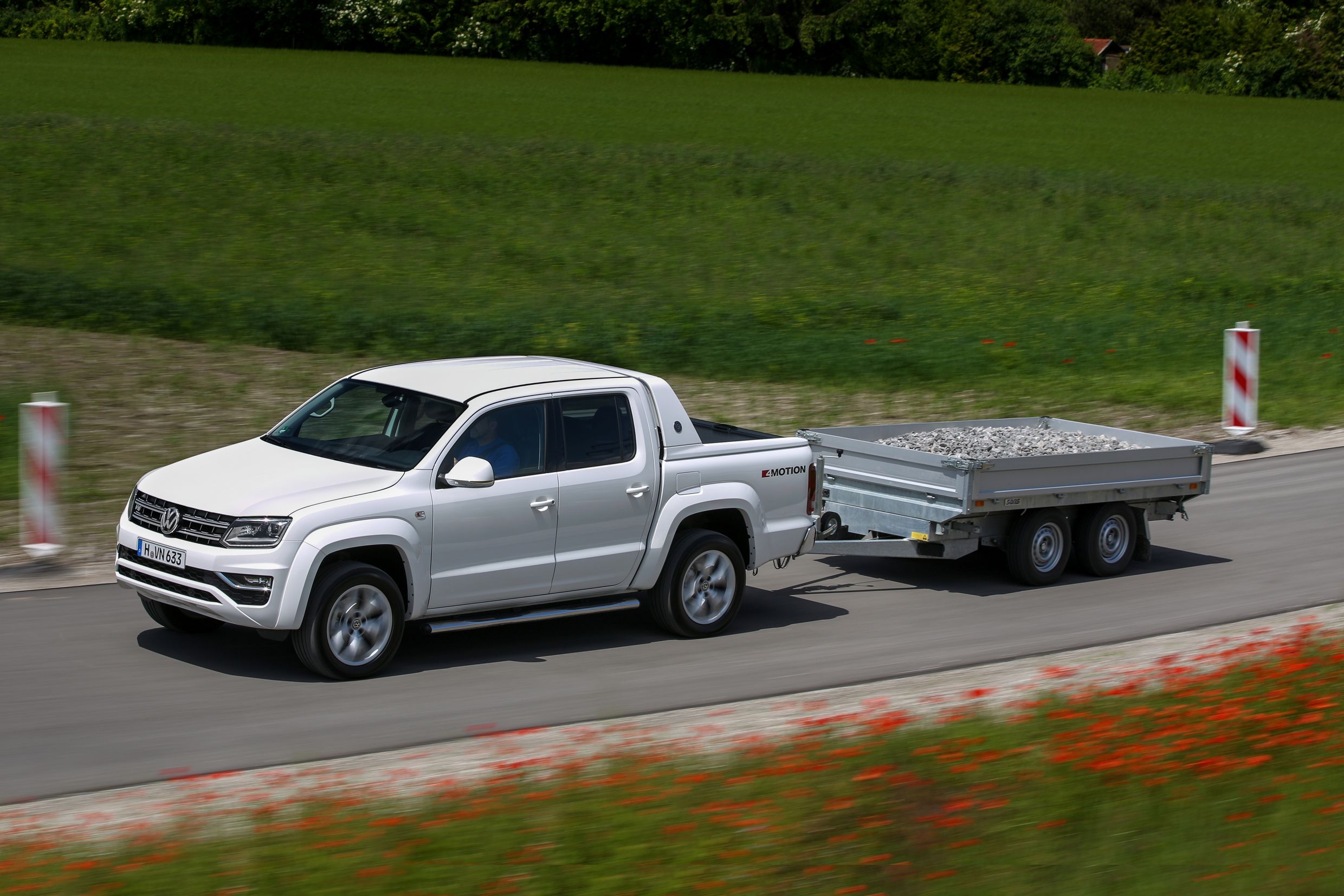 Weißer Pickup mit Anhänger voller Steine fährt auf einer Landstraße durch eine grüne Landschaft unter blauem Himmel.