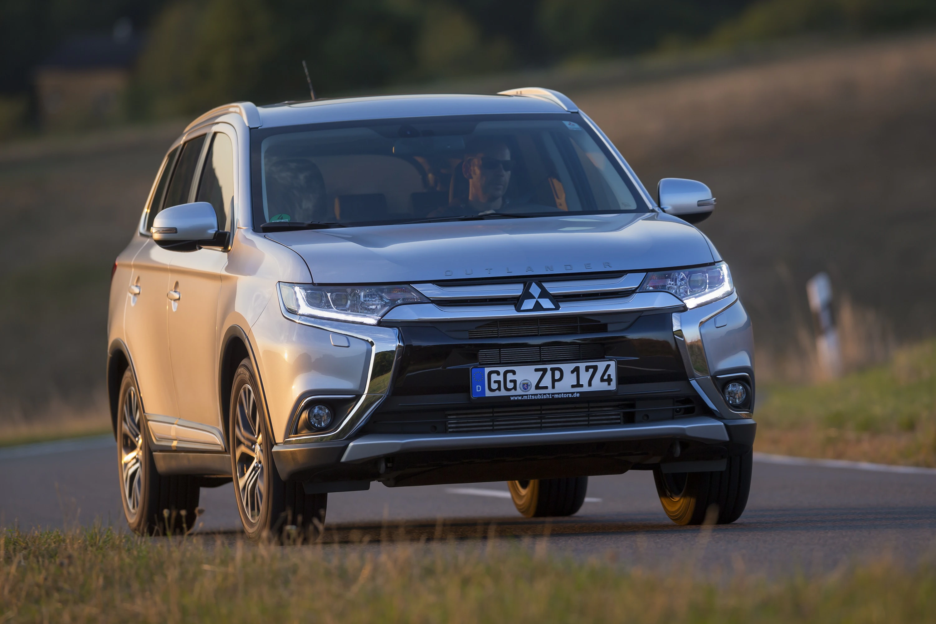 Silberner Mitsubishi SUV fährt auf einer Landstraße bei Tageslicht. Im Hintergrund ist unscharf eine Wiesenlandschaft zu sehen.