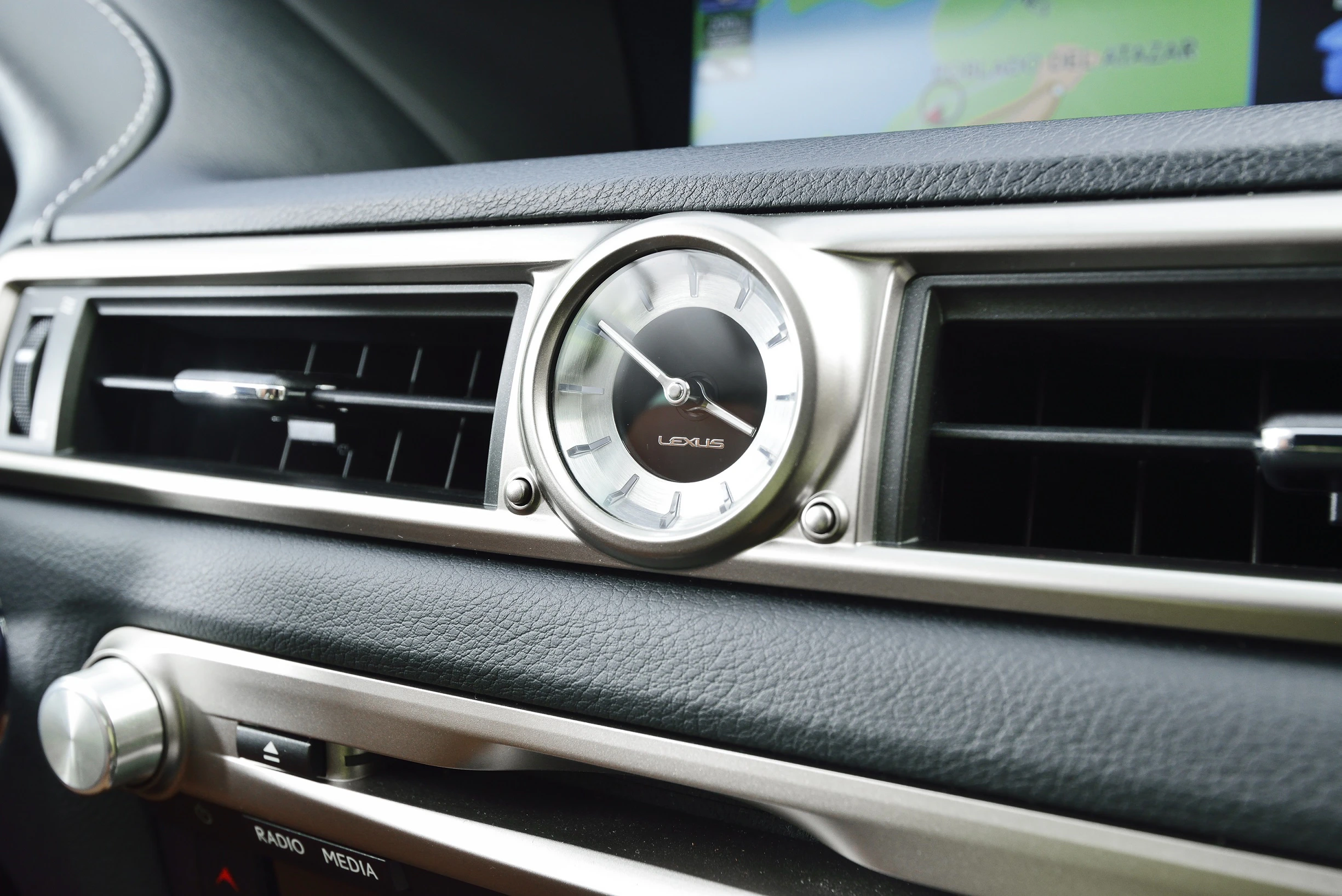Nahaufnahme einer analogen Uhr in einem Autoarmaturenbrett, mit einem Navigationsbildschirm im Hintergrund.