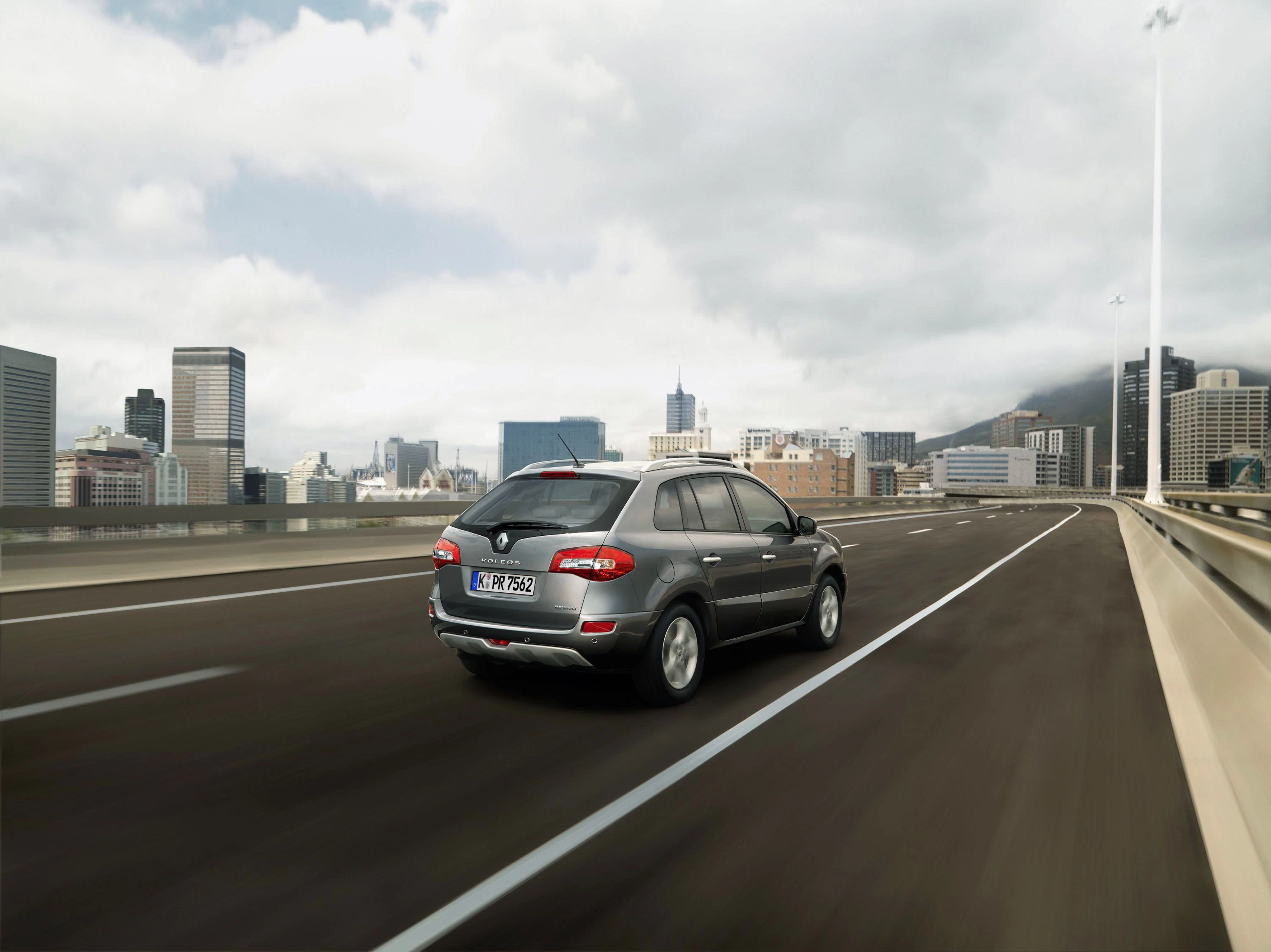 Ein graues Auto fährt auf einer leeren Autobahn mit einer modernen Stadt im Hintergrund unter bewölktem Himmel.