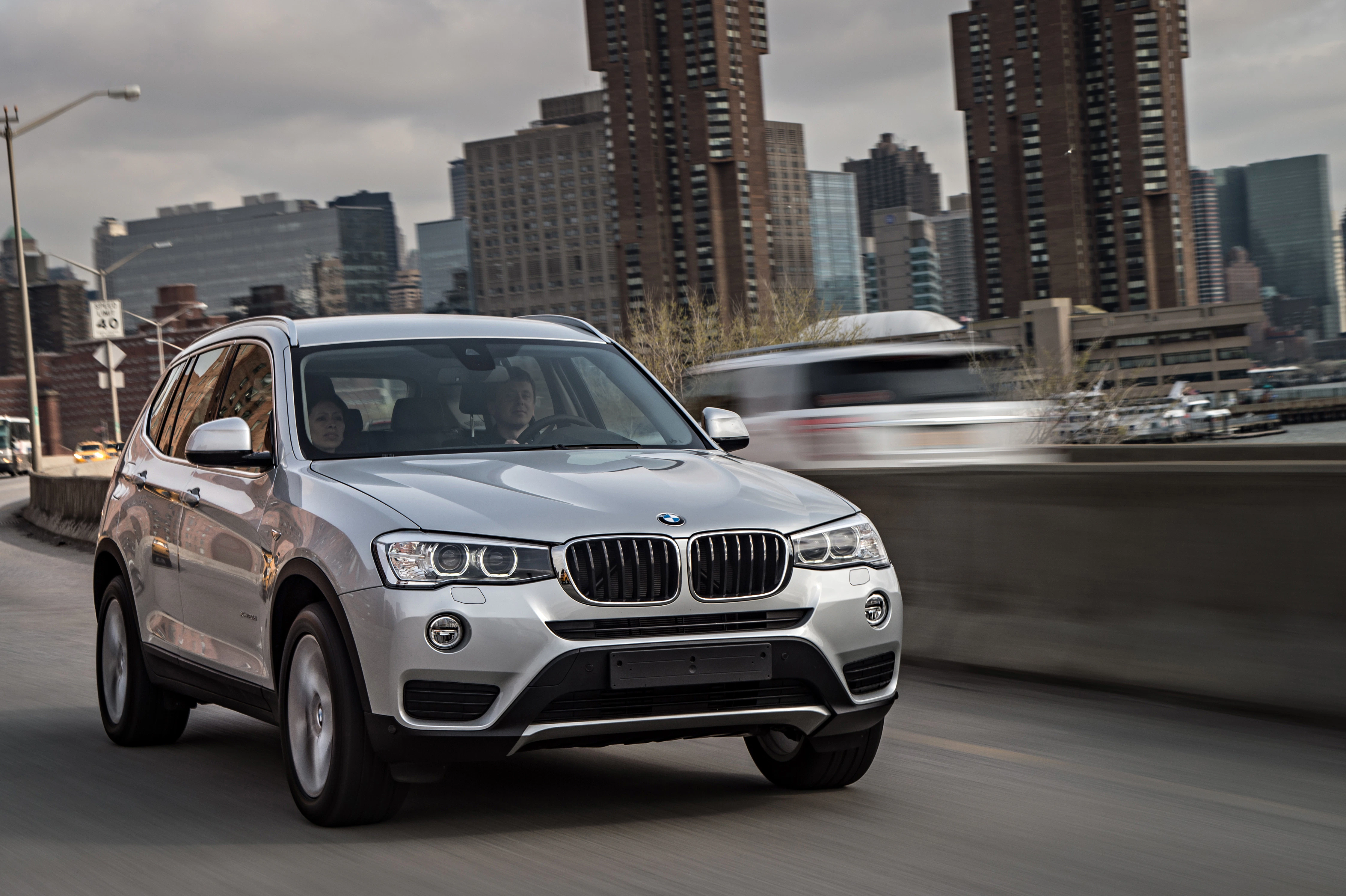 Silberner BMW-SUV fährt auf einer Stadtautobahn bei bewölktem Himmel, hohe Gebäude im Hintergrund.