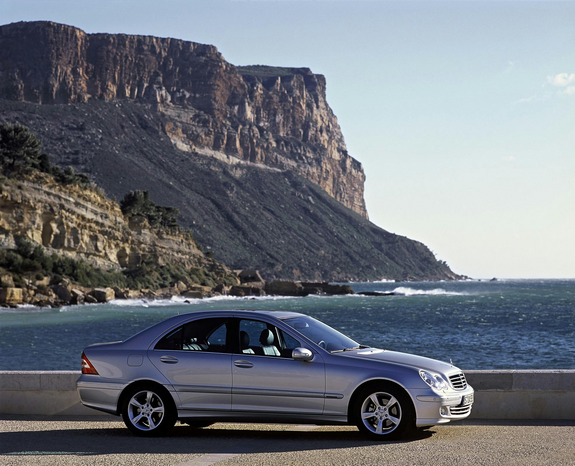 Silberner Mercedes-Benz parkt vor einer Küstenlandschaft mit Felsen und blauem Meer unter klarem Himmel.