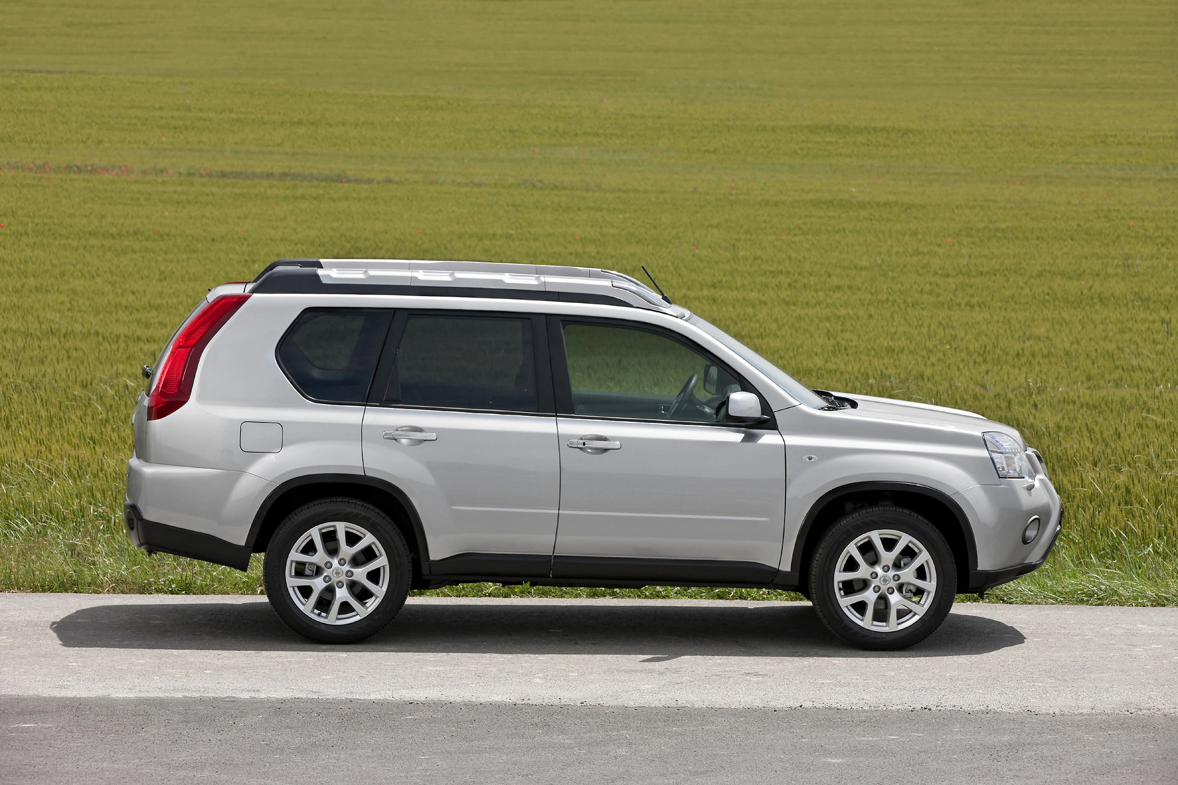 Silberner SUV, seitlich gesehen, steht auf einer Straße vor einem grünen Feld. Klarer Himmel im Hintergrund.