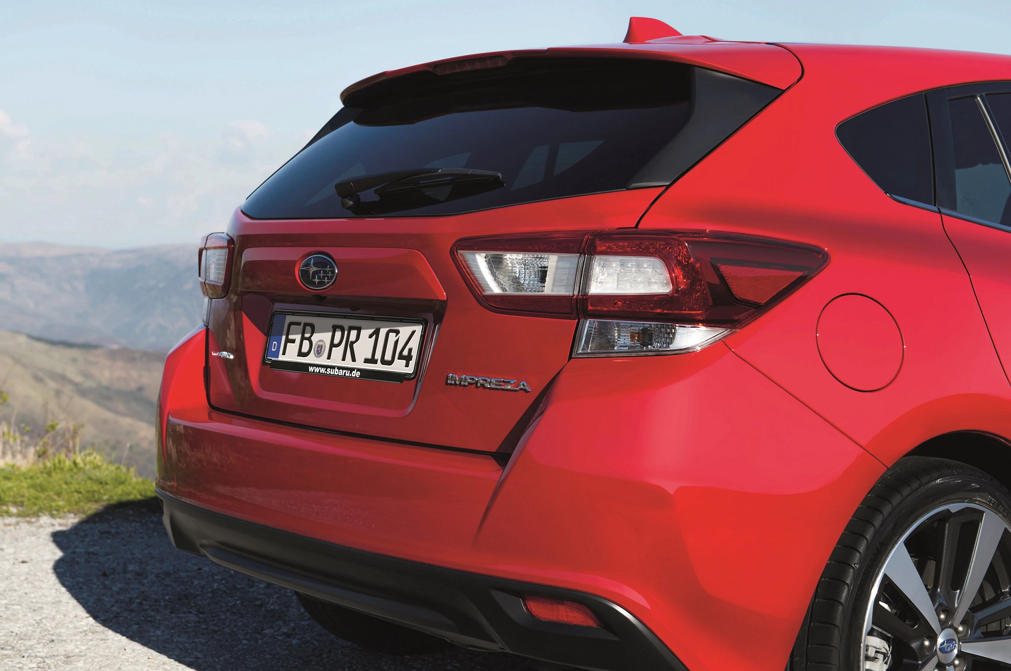 Rotes Auto (Subaru Impreza) von hinten, parkt auf einem Hügel mit Bergblick. Deutsches Kennzeichen sichtbar.