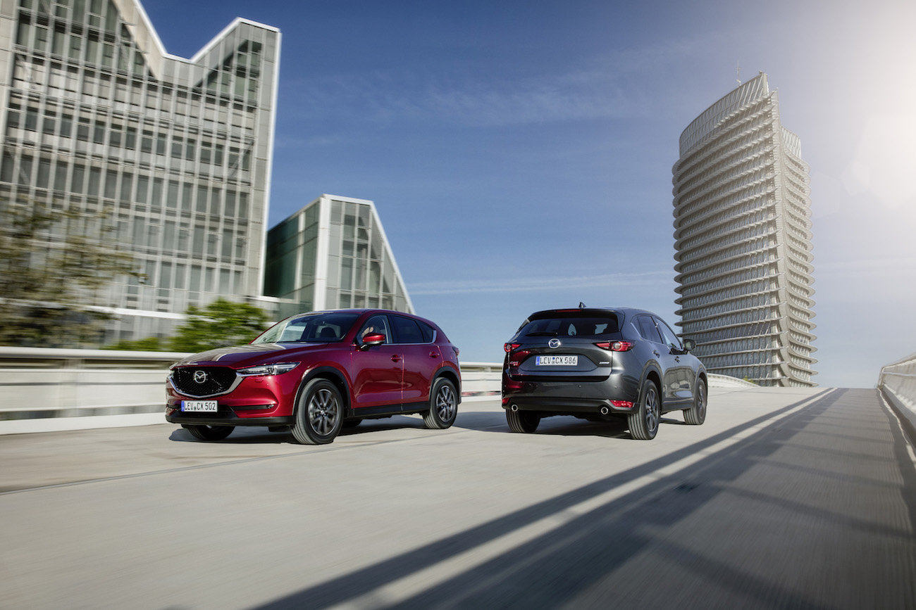 Zwei Mazda CX-5 SUVs fahren auf einer modernen Stadtautobahn mit futuristischen Gebäuden im Hintergrund unter blauem Himmel.