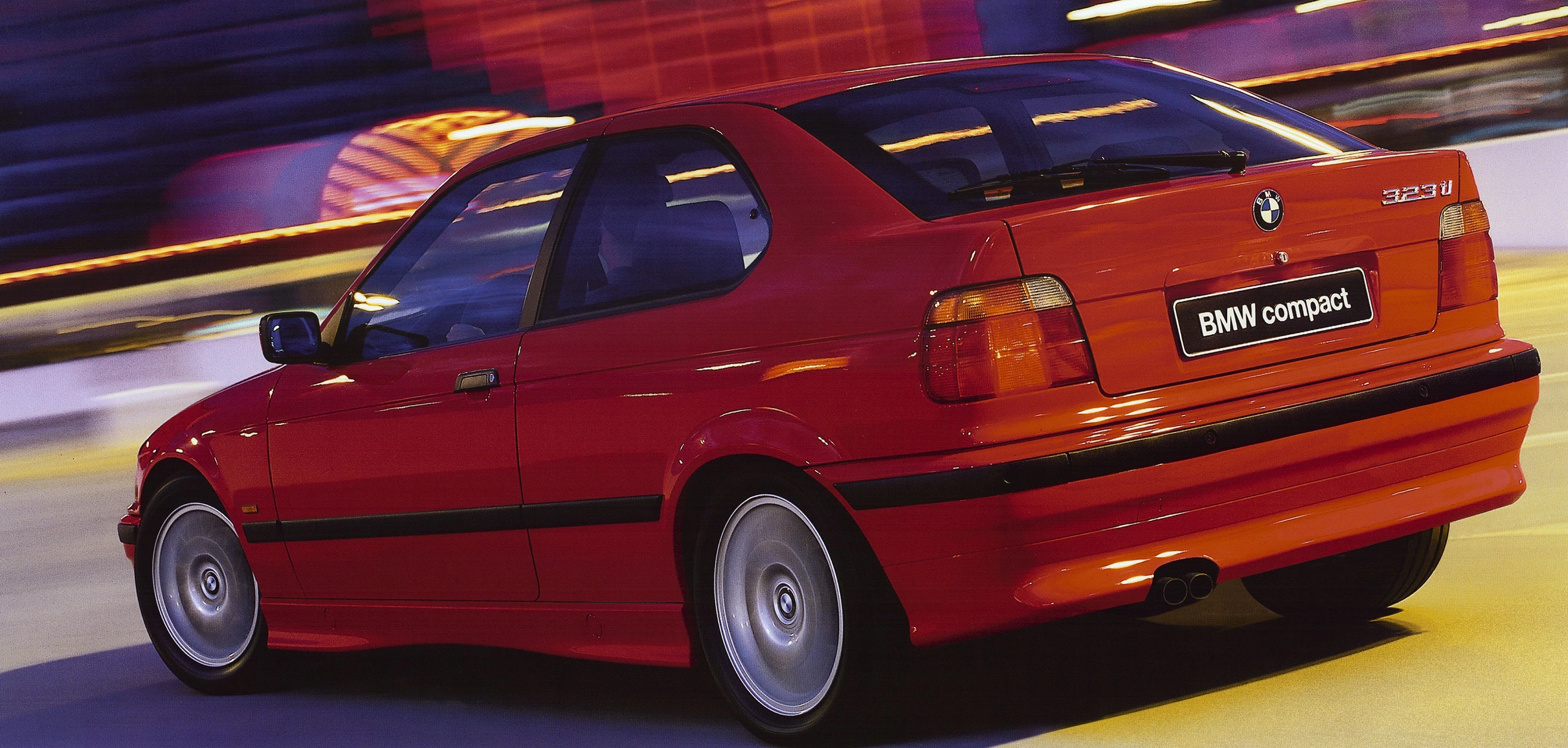Roter BMW 323ti Compact von hinten, fährt durch eine dynamisch verschwommene Stadtumgebung.