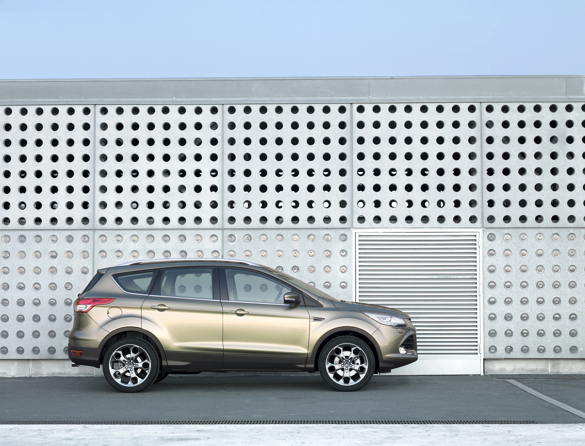 SUV in goldener Farbe vor einer modernen Fassade mit runden Löchern, auf einem grauen Parkplatz abgestellt.