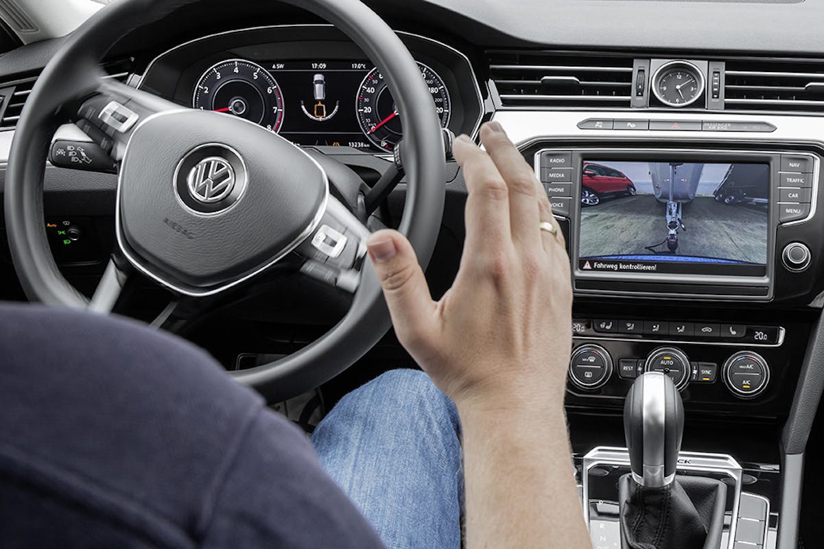 Fahrer steuert Auto mit Rückfahrkamera-Anzeige auf dem Bildschirm. Hands-free-Funktion im Einsatz. Lenkrad mit VW-Logo sichtbar.