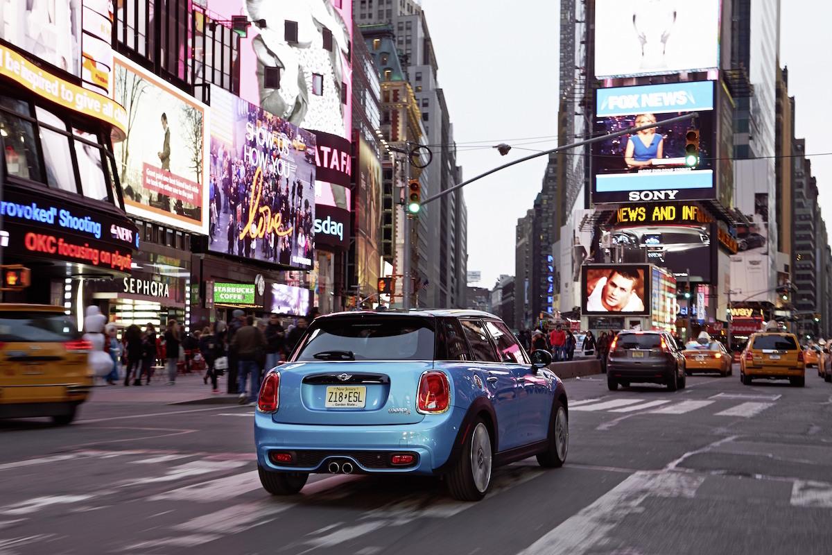 Blauer Mini Cooper fährt durch eine belebte Straße mit leuchtenden Werbetafeln und gelben Taxis bei Abenddämmerung.